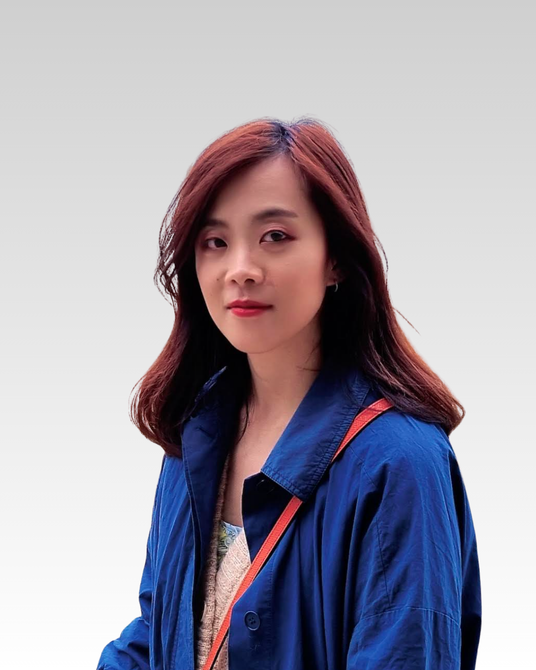 Una donna con i capelli castani e rossicci, che indossa una giacca blu e una borsa arancione, si trova contro uno sfondo grigio chiaro.