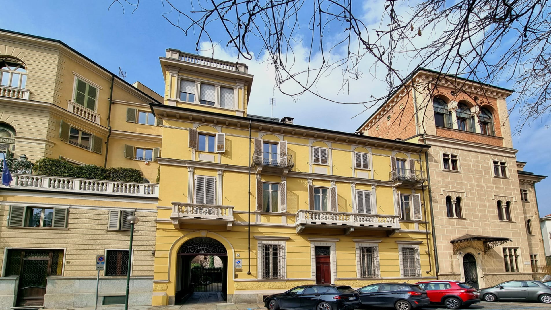 Edificio storico con facciata gialla e dettagli in pietra, finestre con persiane, balconi in ferro battuto e un arco di ingresso, con auto parcheggiate davanti.