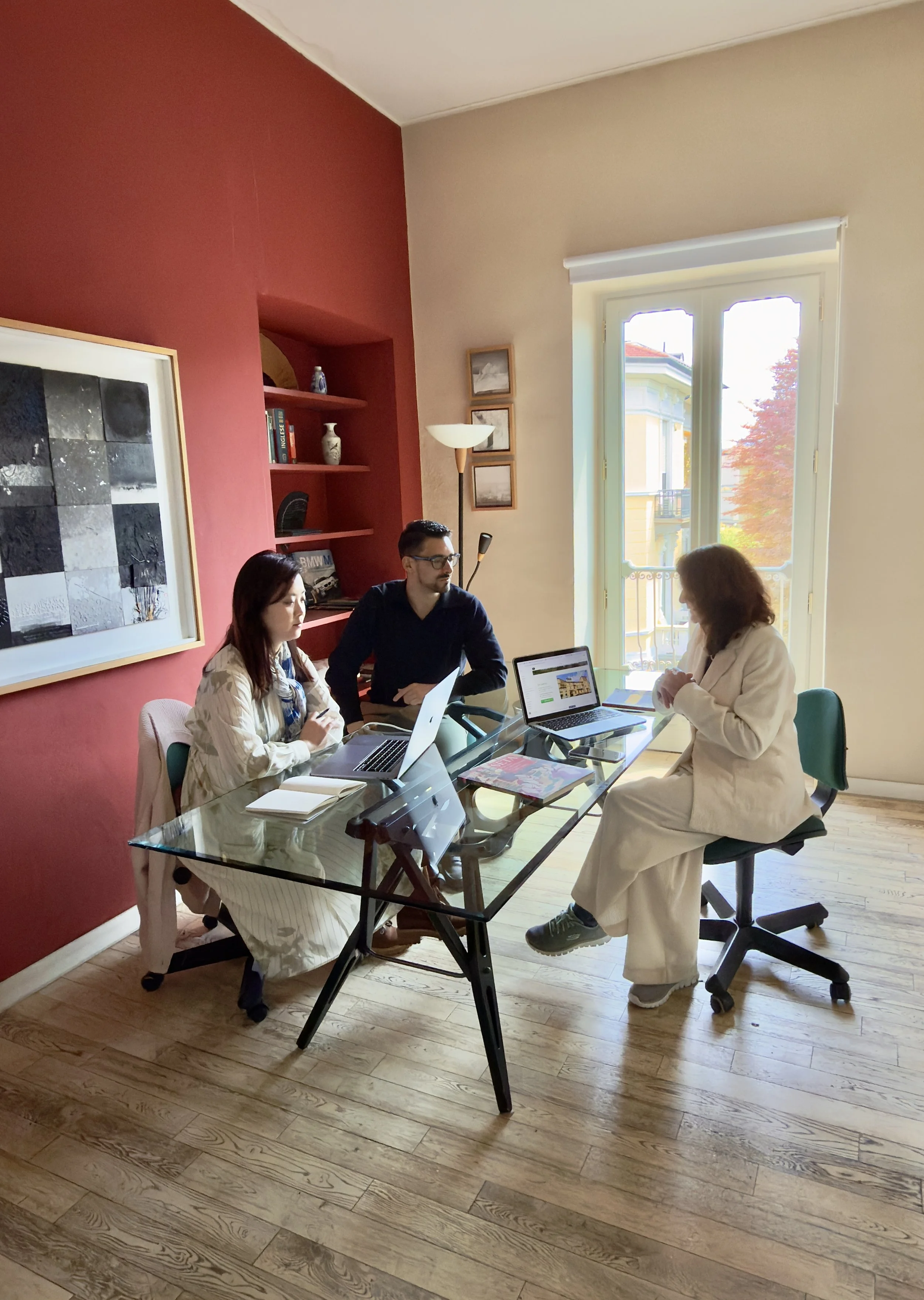 Tre persone sedute intorno a un tavolo di vetro in una stanza luminosa mentre discutono, con laptop e documenti sul tavolo.