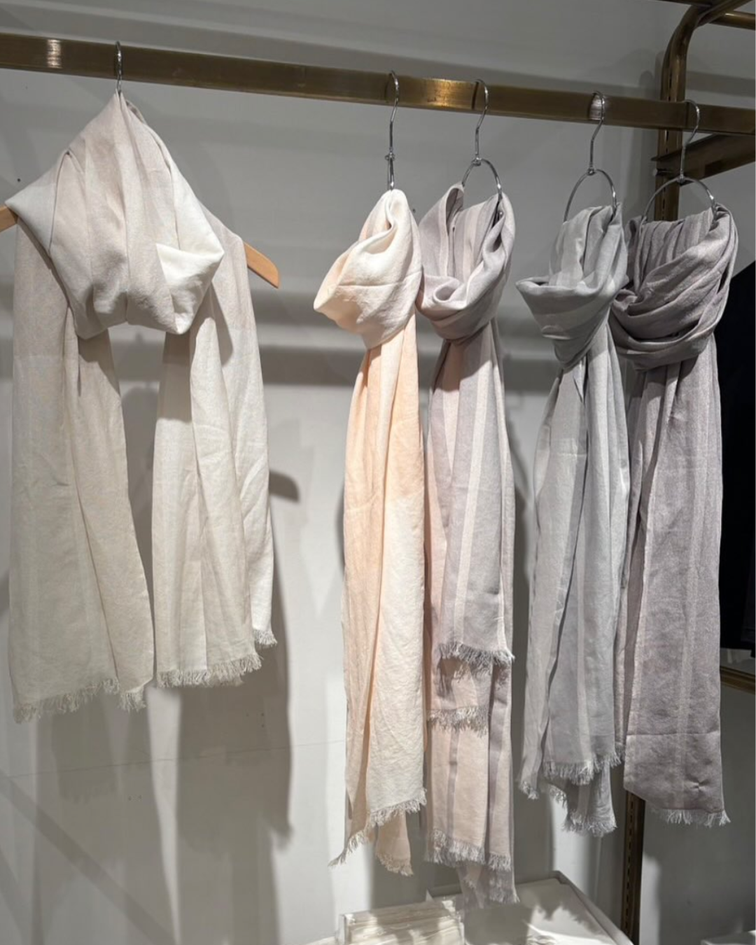 Quattro sciarpe leggere di diversi colori pastello appese a un bastone con ganci, in un negozio di abbigliamento.