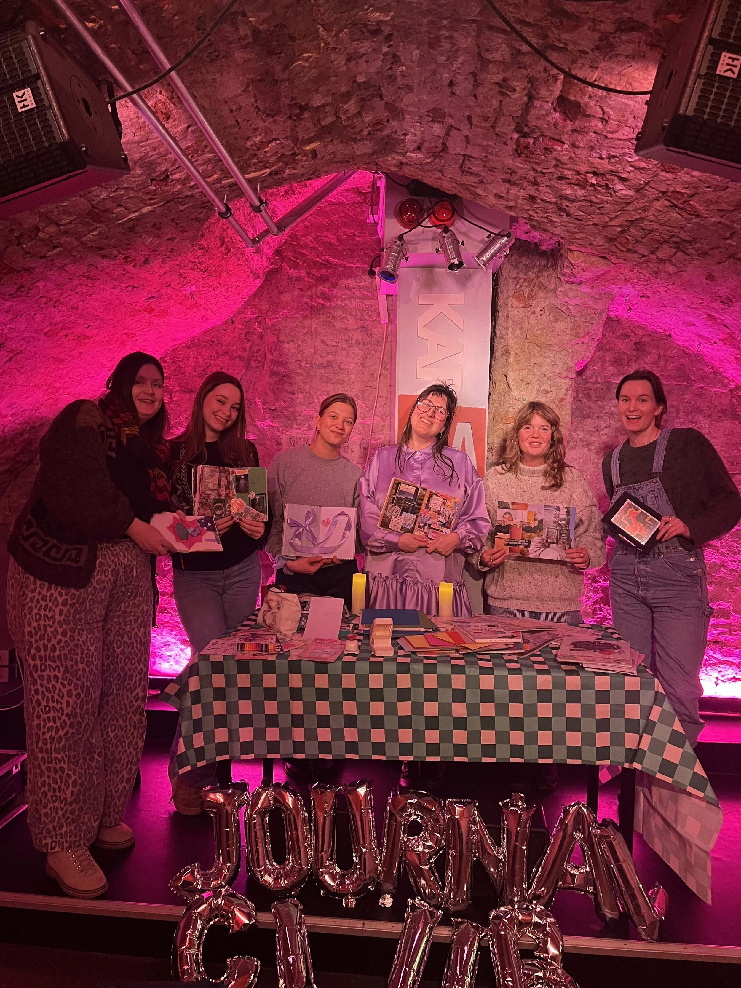 Zes vrouwen staan achter een tafel vol verschillende gepresenteerde handgemaakte creaties, met een bakstenen muur en pinkverlichte achtergrond. De tafel heeft een zwart-wit geruite tafelkleed en er hangen metallic ballonnen die 'JOURNAL CLUB' vormen.