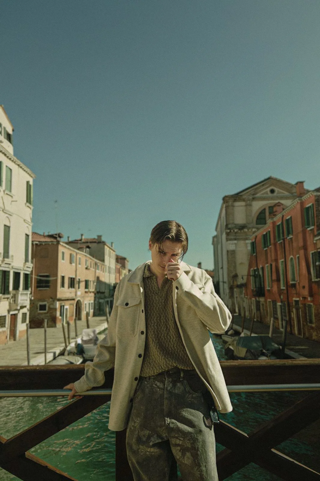 Young adult with short hair wearing a beige jacket and camouflage pants standing on a bridge in Venice, Italy, with colorful buildings and boats in the background on a sunny day.