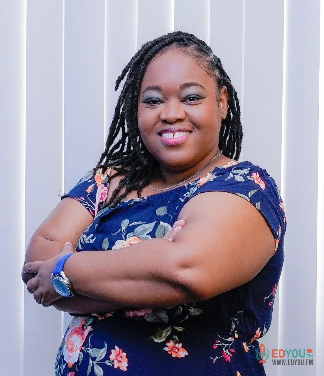 A woman with dark skin and black dreadlocks smiling with arms crossed, wearing a floral blue dress, standing in front of a white background with vertical blinds.
