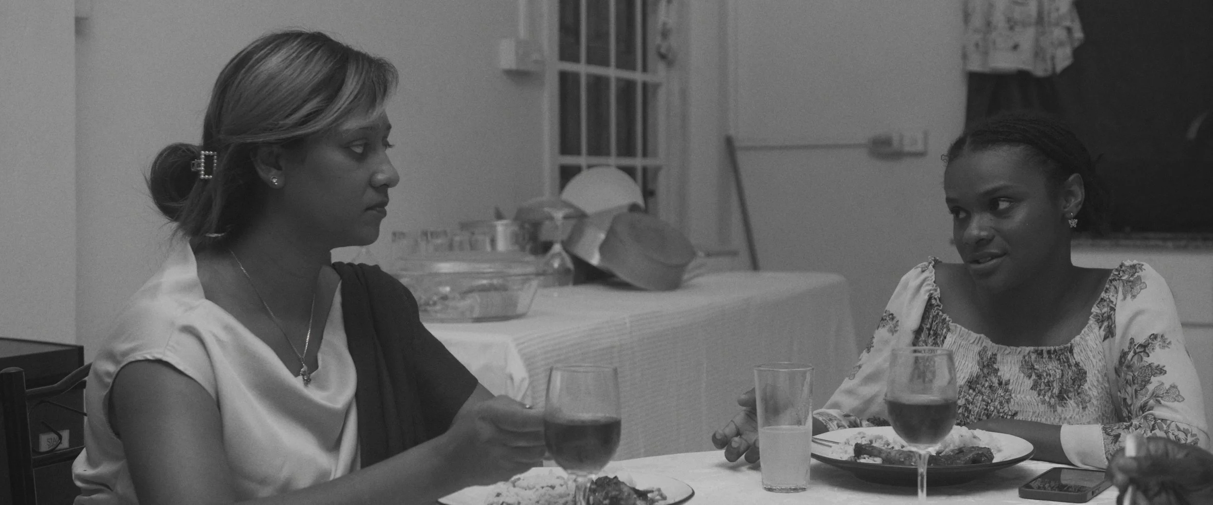 Two women sitting at a table having a conversation, with glasses of wine and plates of food in front of them, in a black-and-white photo.