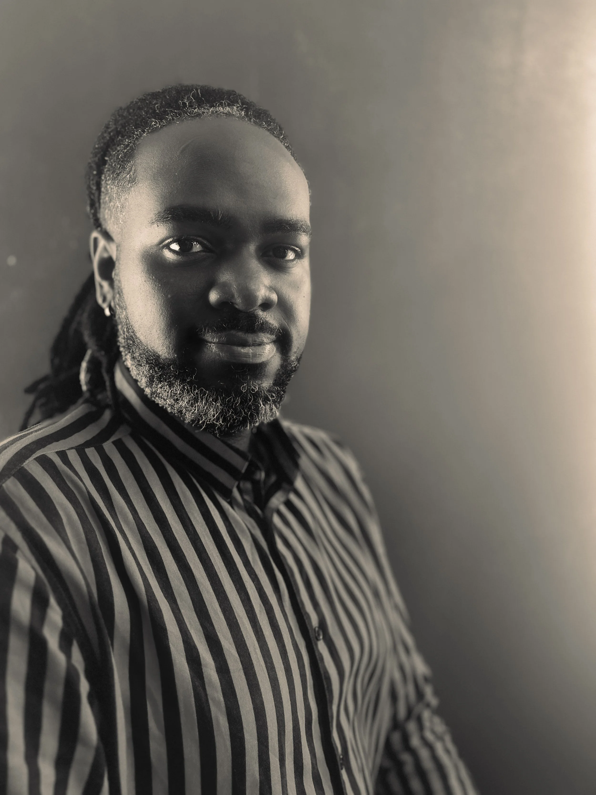 Black and white portrait of a man with dreadlocks, beard, and earrings, wearing a striped shirt, standing against a plain background.