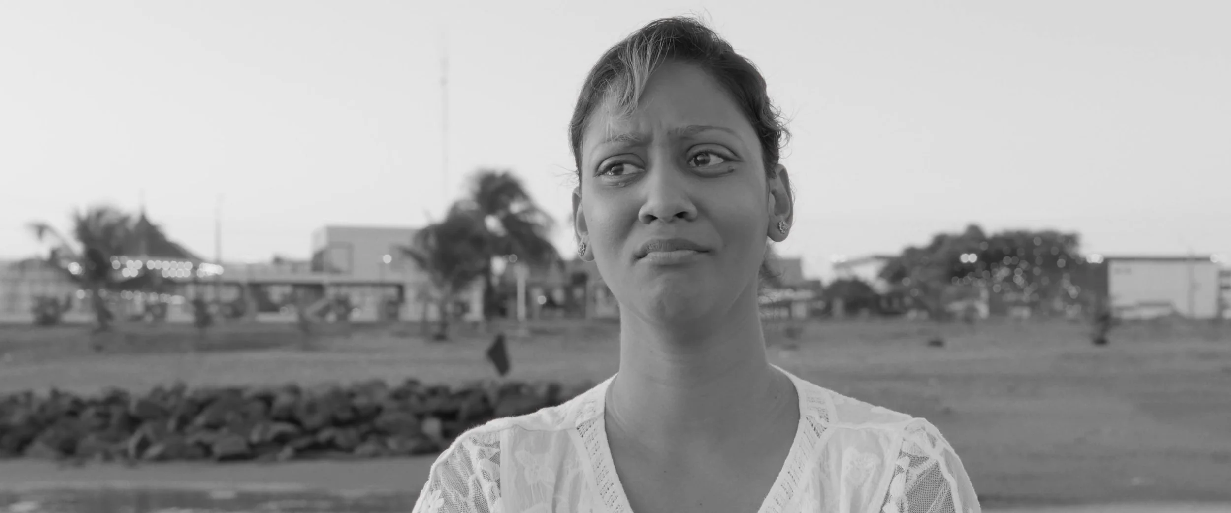 Black and white image of a woman with a puzzled or worried expression, standing outdoors with trees and buildings in the background.