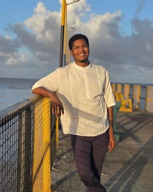 A smiling young man leaning on a yellow railing on a seaside pier during sunset or late afternoon, with ocean waves and cloudy sky in the background.