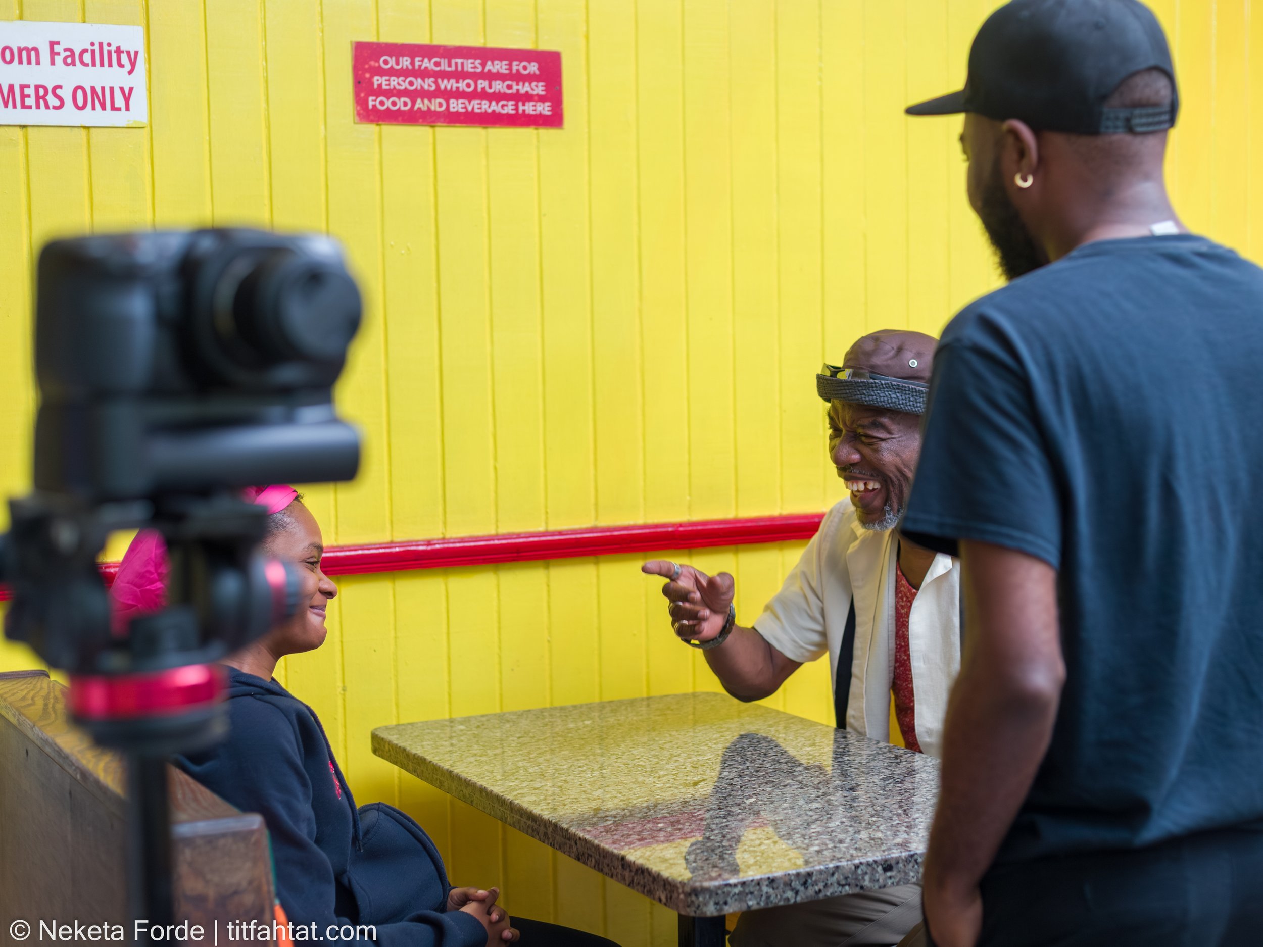 Three people in conversation inside a restaurant with bright yellow walls. One woman is seated, smiling and wearing a pink headscarf. Two men are standing and sitting, one with a beard, cap, and earrings, the other wearing a hat and sunglasses, all engaged in a lively chat.