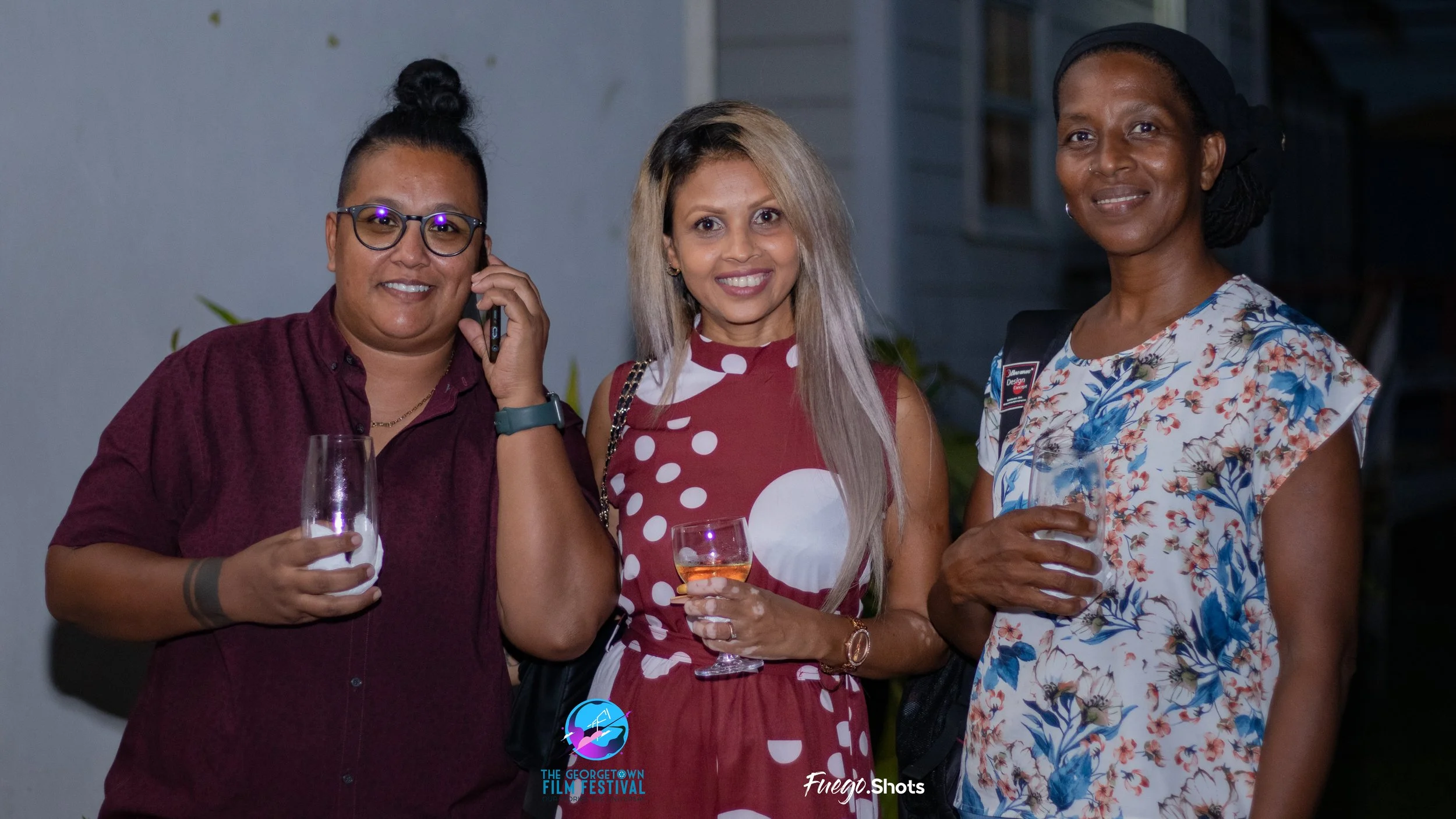 Three women at an outdoor event, smiling and holding drinks, with one woman talking on a cell phone. The woman on the left wears glasses and a dark red shirt, the woman in the middle wears a red dress with white polka dots, and the woman on the right