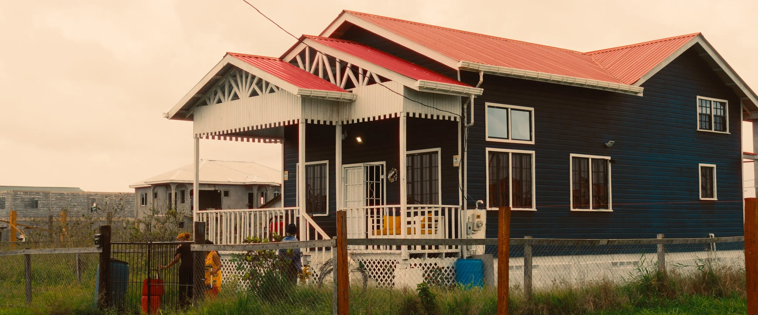 A two-story house with a dark blue exterior, white trim, and a red metal roof, featuring a front porch with white railings and columns, surrounded by a grassy yard enclosed with a chain-link fence. There are two children in the yard, one holding a garden tool, and the other standing near a bicycle.