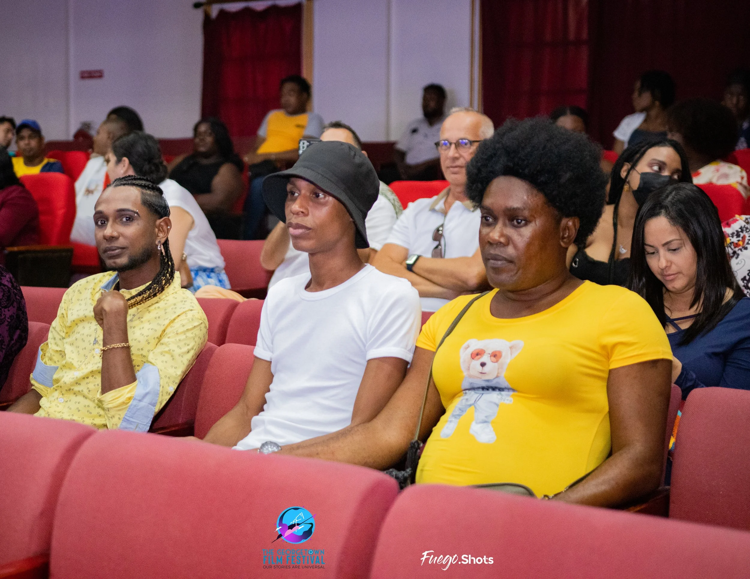 Audience seated in red theater seats during an event, with a diverse group of people paying attention, including a woman in yellow with a graphic of a bear on her T-shirt, and other attendees in the background.
