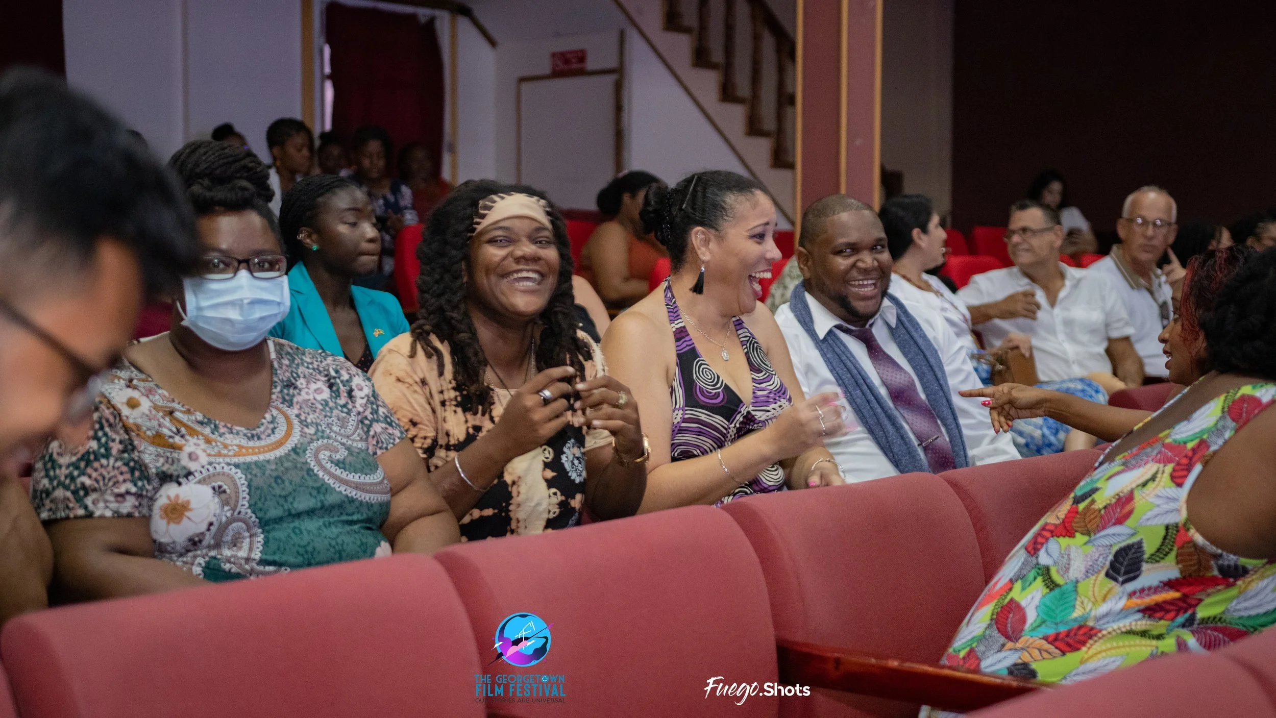 People sitting in an auditorium laughing and smiling, including four women in the front row and other audience members in the background, some wearing masks.