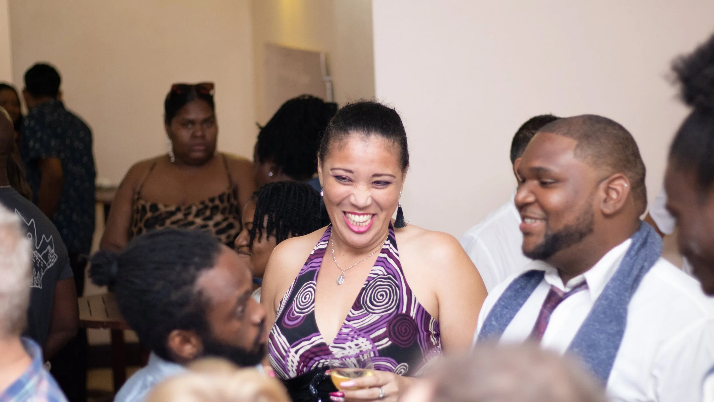 Group of people socializing indoors, with a smiling woman in a purple patterned dress at the center holding a drink, and a man in a white shirt and vest smiling beside her.