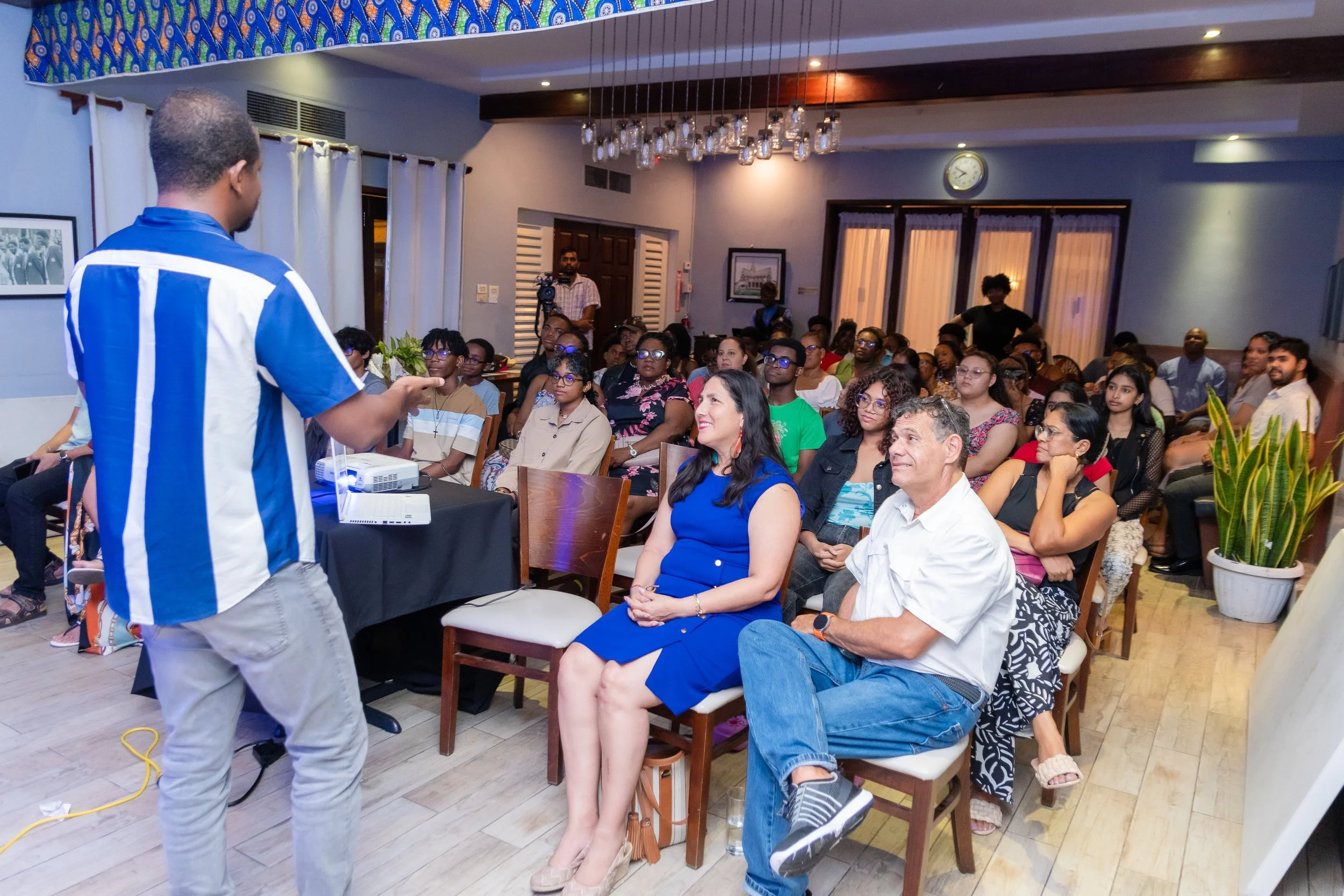 Man in blue and white striped shirt giving a presentation in a room full of seated audience members, some smiling and attentive.