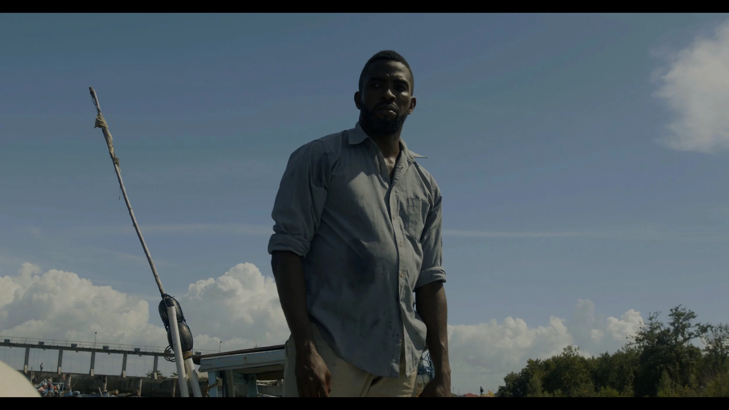 A man with a beard and dark skin, wearing a light-colored, rolled-up shirt, standing outdoors under a cloudy sky, with a boat and trees in the background.