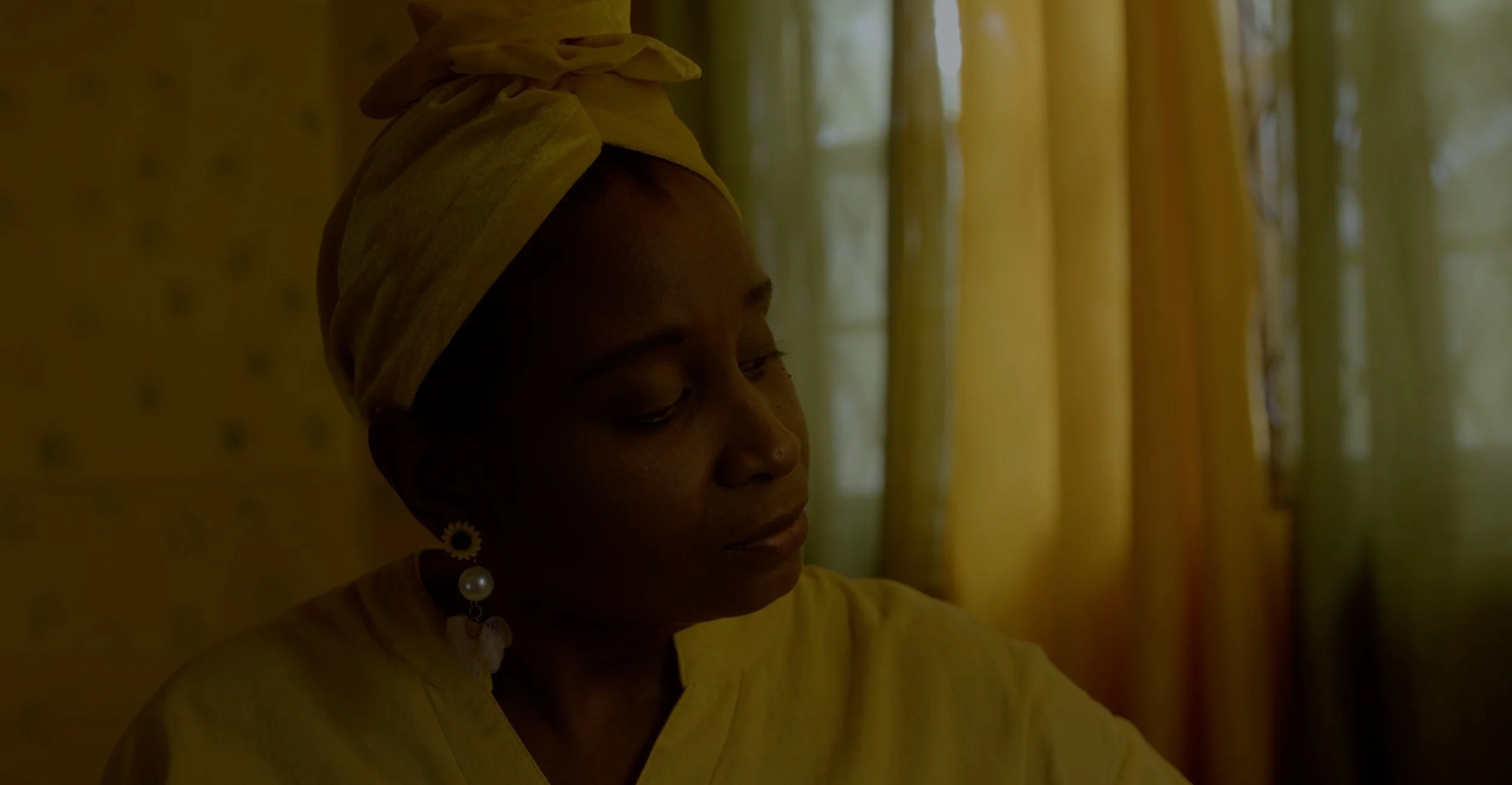 A woman with dark skin wearing a yellow headwrap and yellow clothing, looking down with a calm expression, standing in a room with yellow curtains and a patterned yellow wall.