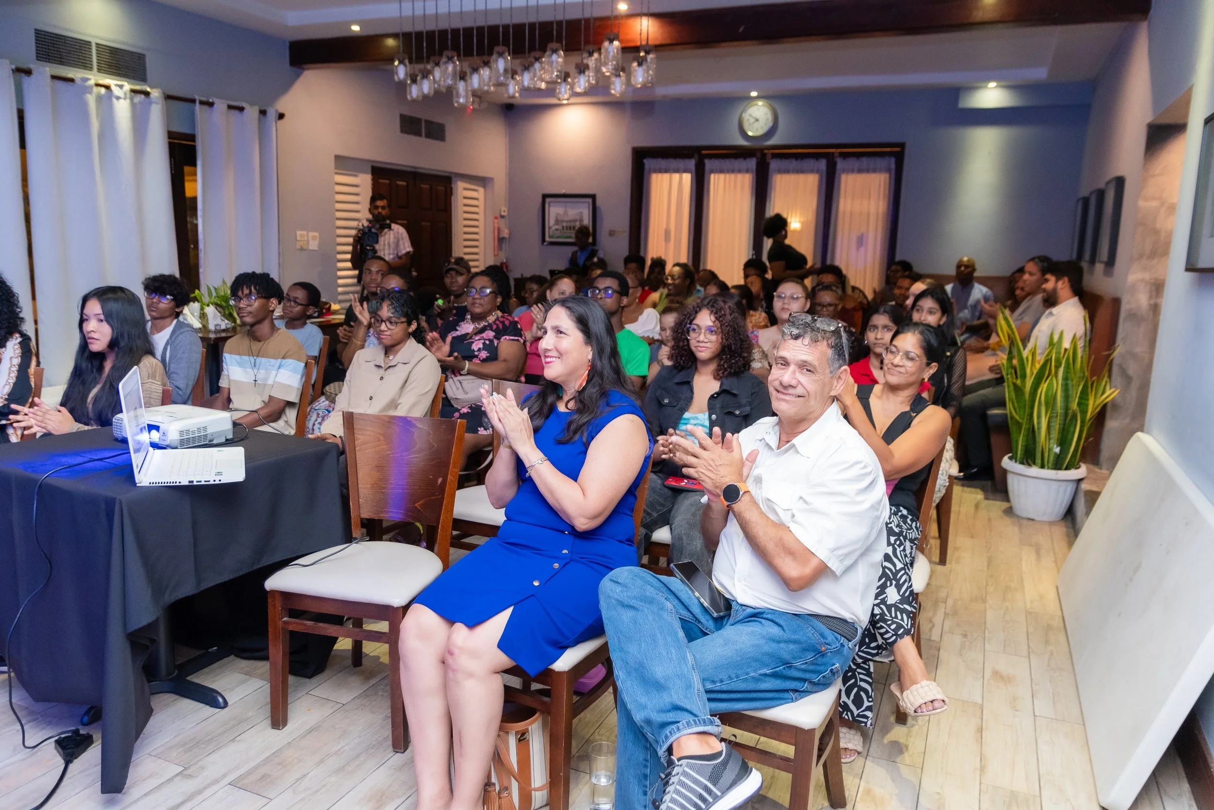An indoor event with an audience of diverse people sitting and clapping, with some smiling, in a well-lit room with curtains, potted plants, and framed pictures on the walls.