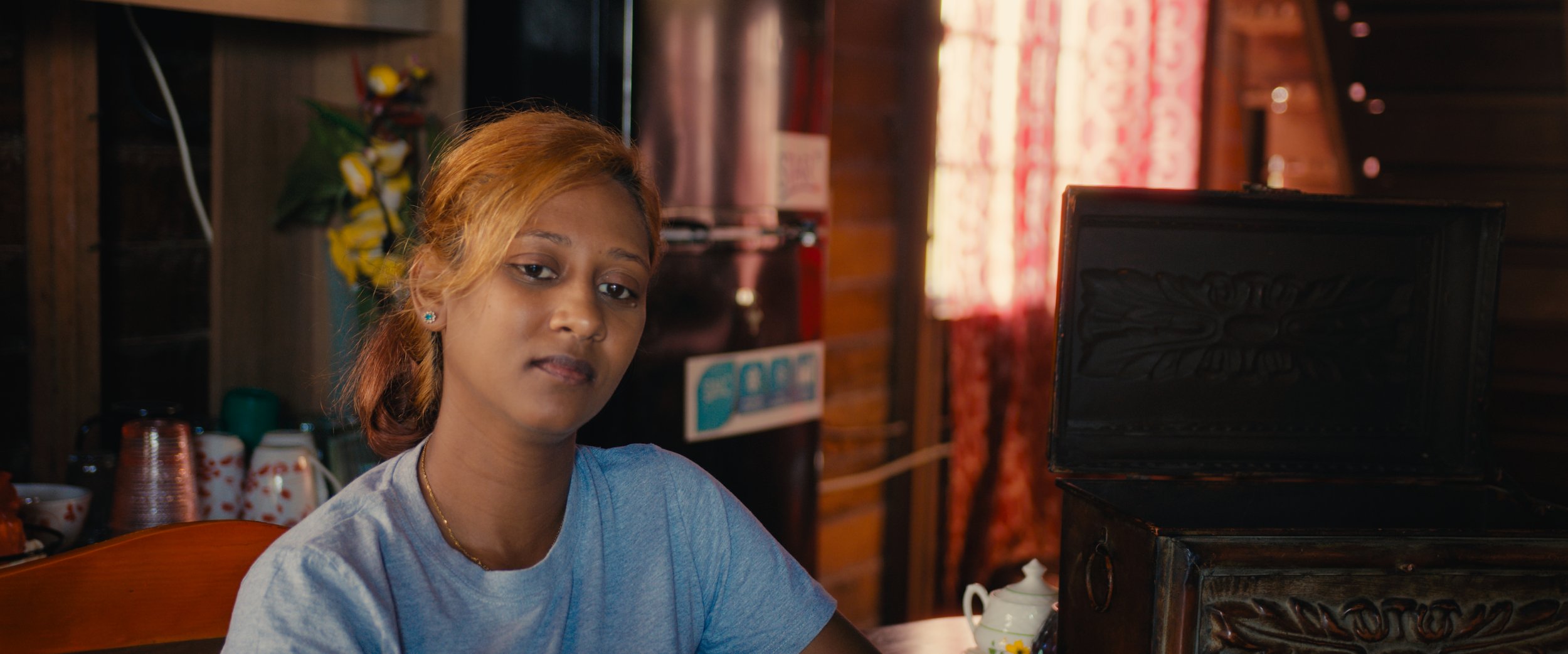 A young woman with light brown hair, wearing a light gray T-shirt and blue earrings, sitting at a wooden table in a cozy room with wood-paneled walls, a teapot, cups, and a black, open jewelry box on the table.