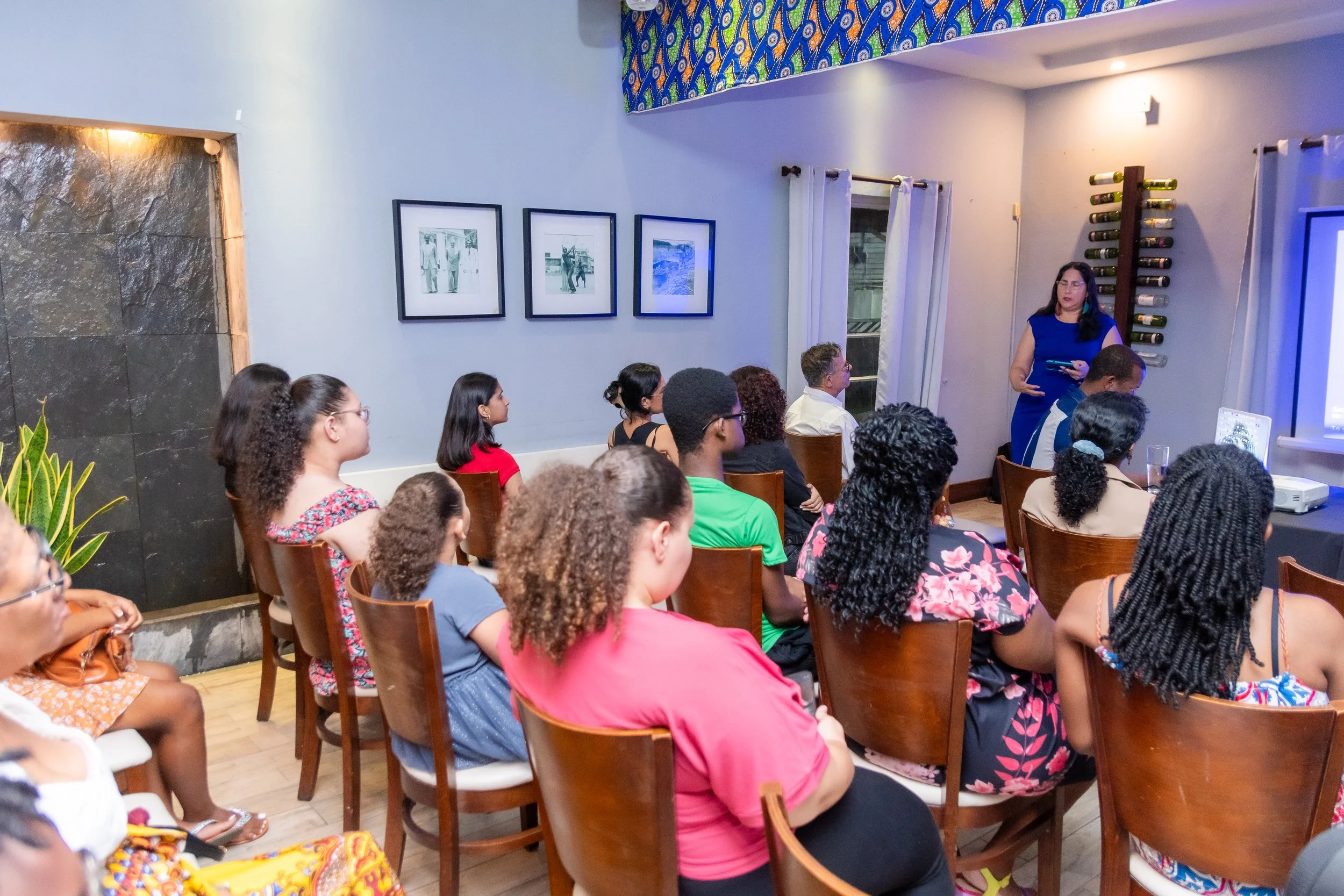 A group of people seated in a room watching a woman giving a presentation at the front. The room has framed photos on the wall, a patterned fabric on the ceiling, and a window with white curtains. There is a wine rack on the wall and a large screen or monitor being used for the presentation.