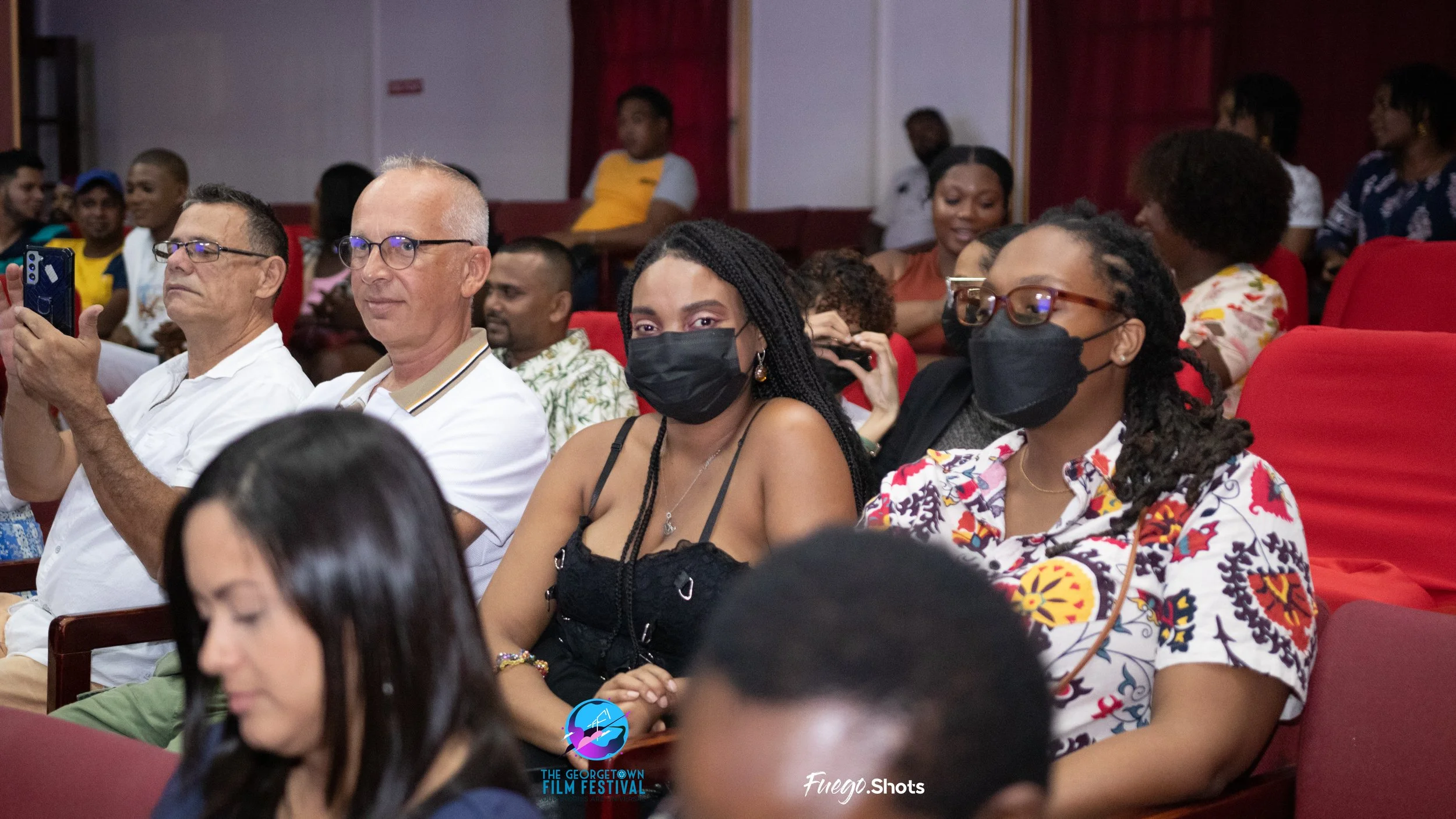 Audience attending a film festival, some wearing black masks and glasses, sitting in red theater seats, with a logo of The Georgetown Film Festival and a photographer's watermark at the bottom.