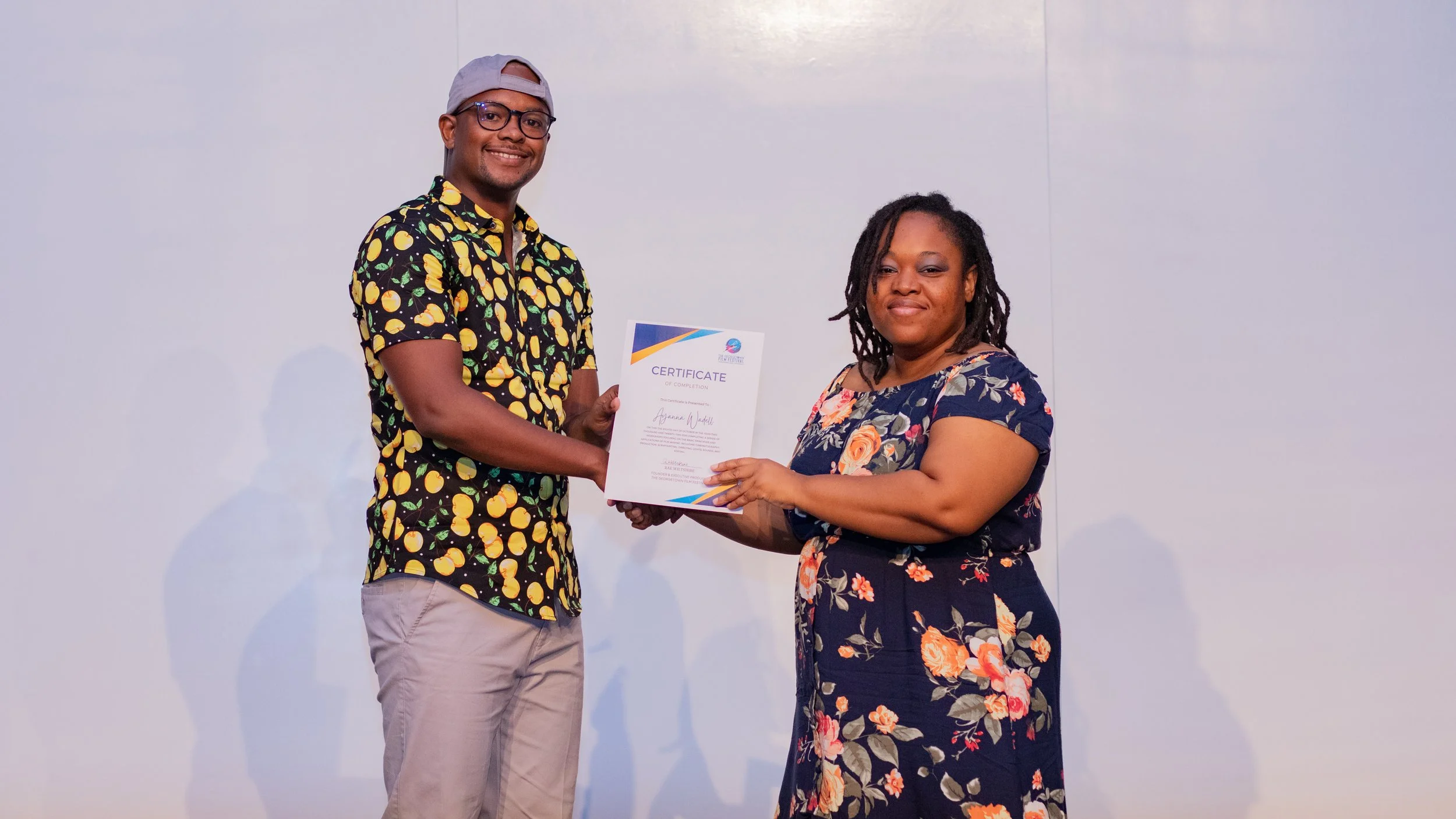 A man wearing glasses and a cap, dressed in a lemon-patterned shirt and beige pants, smiles while receiving a certificate from a woman in a floral dress, standing against a plain white wall.