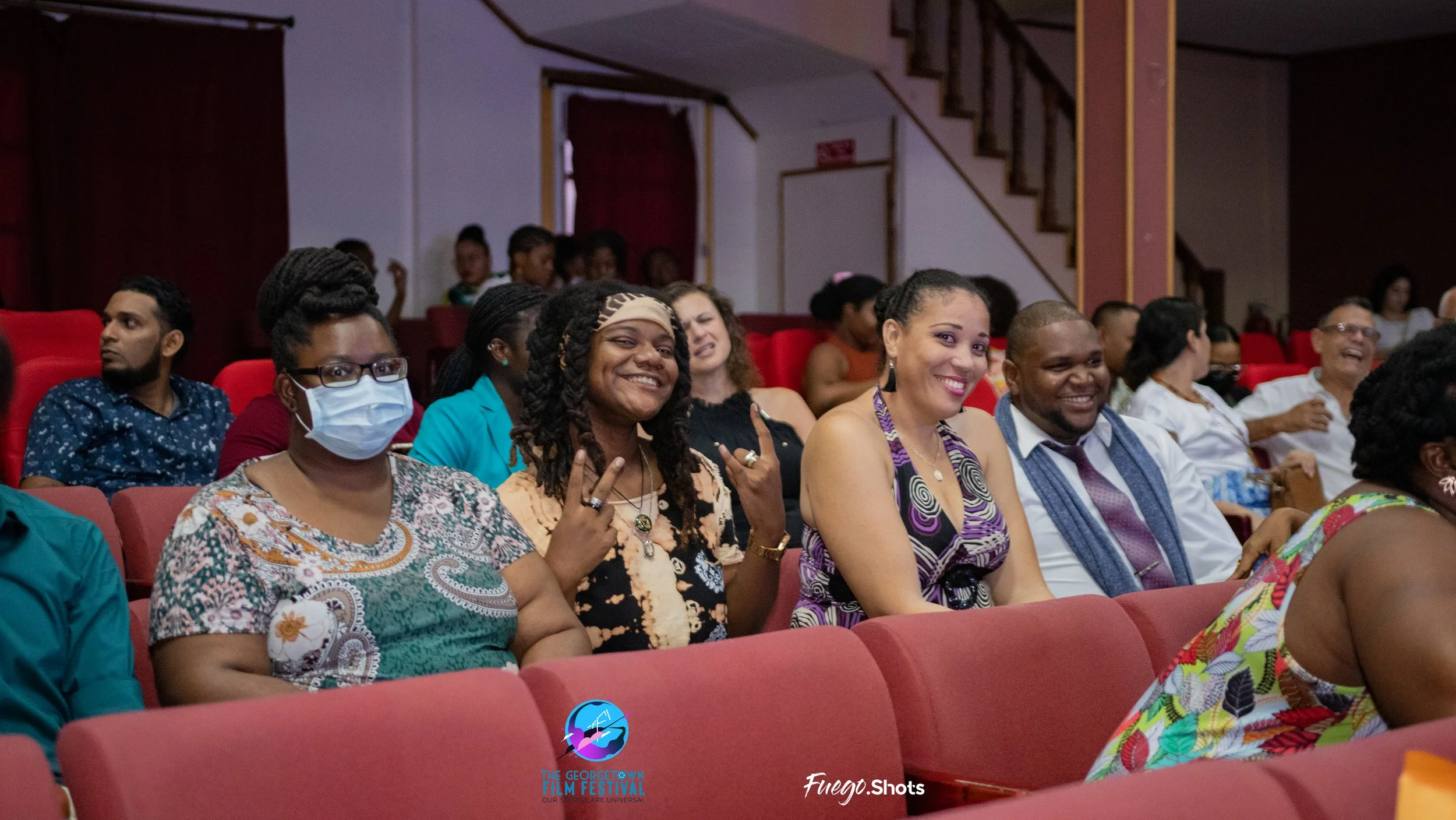 Audience sitting in red theater chairs at a film festival, smiling and enjoying the event. Some are wearing masks, and audience members are of diverse backgrounds.