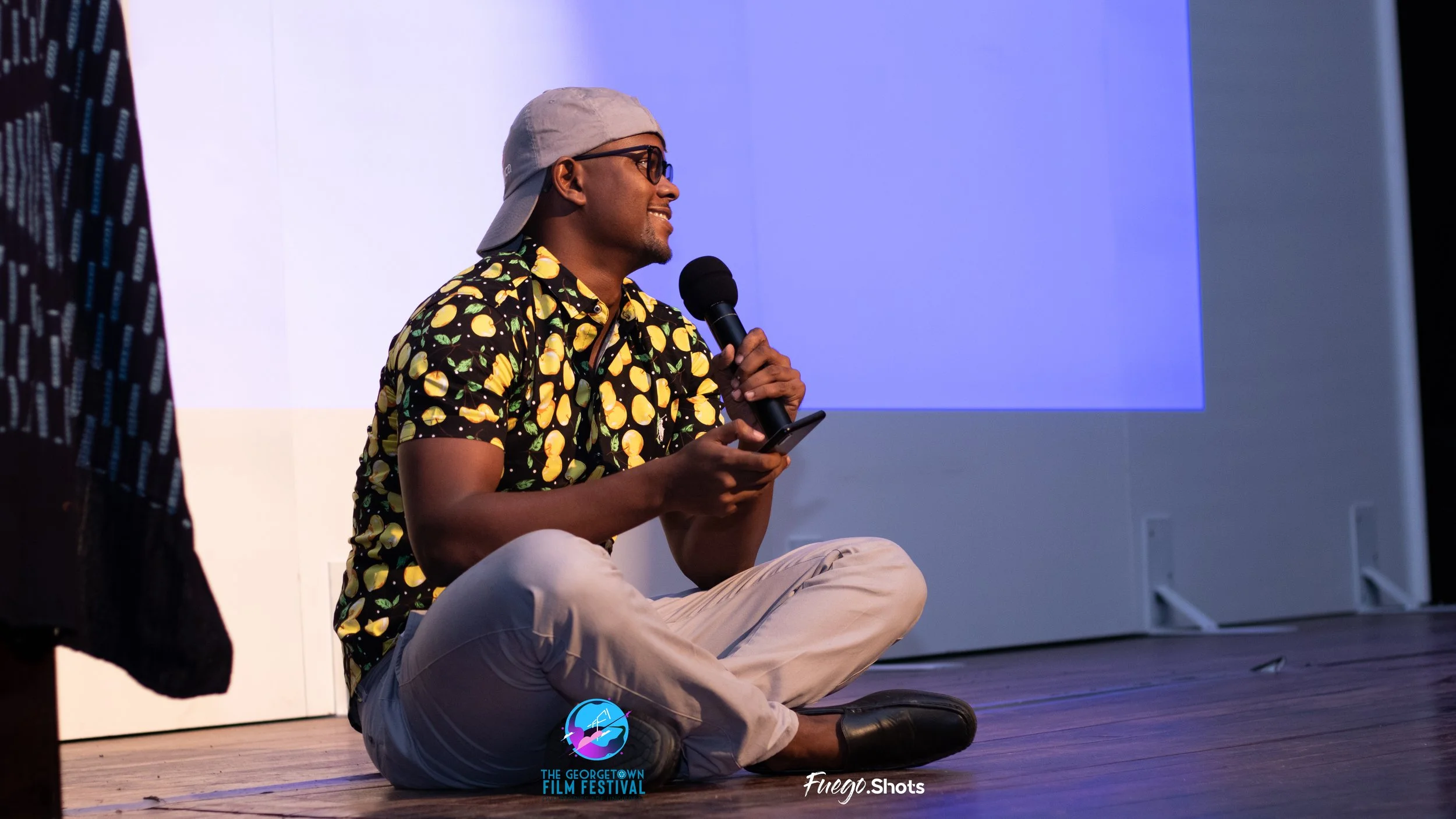 Young man sitting cross-legged on a stage, holding a microphone and smartphone, wearing a lemon-patterned shirt, light-colored pants, a gray cap worn backwards, and glasses, with a projection screen behind him at the Georgetown Film Festival.