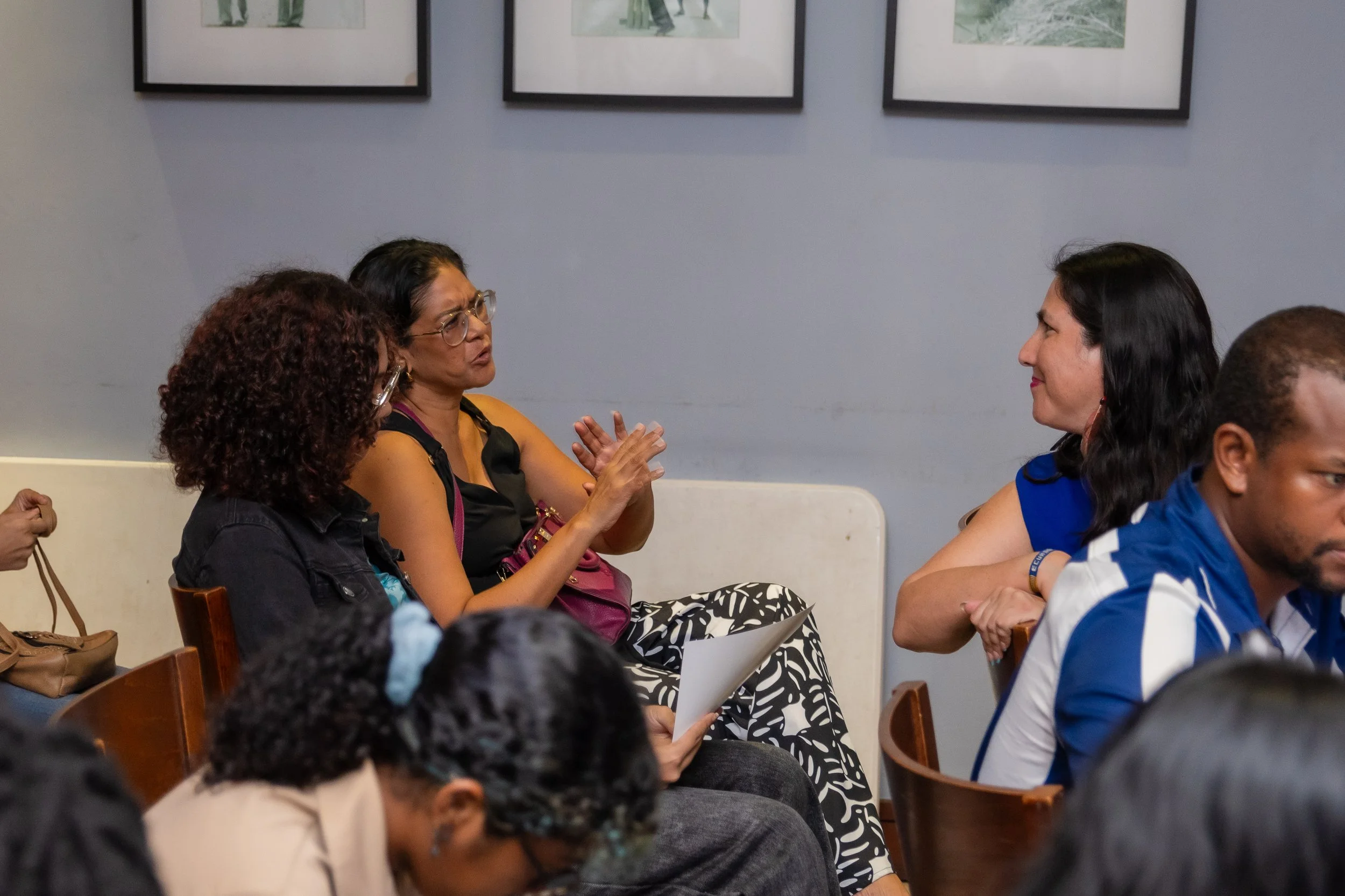 Women engaged in a discussion in a conference room, sitting on chairs with a plain blue wall and framed pictures behind them.