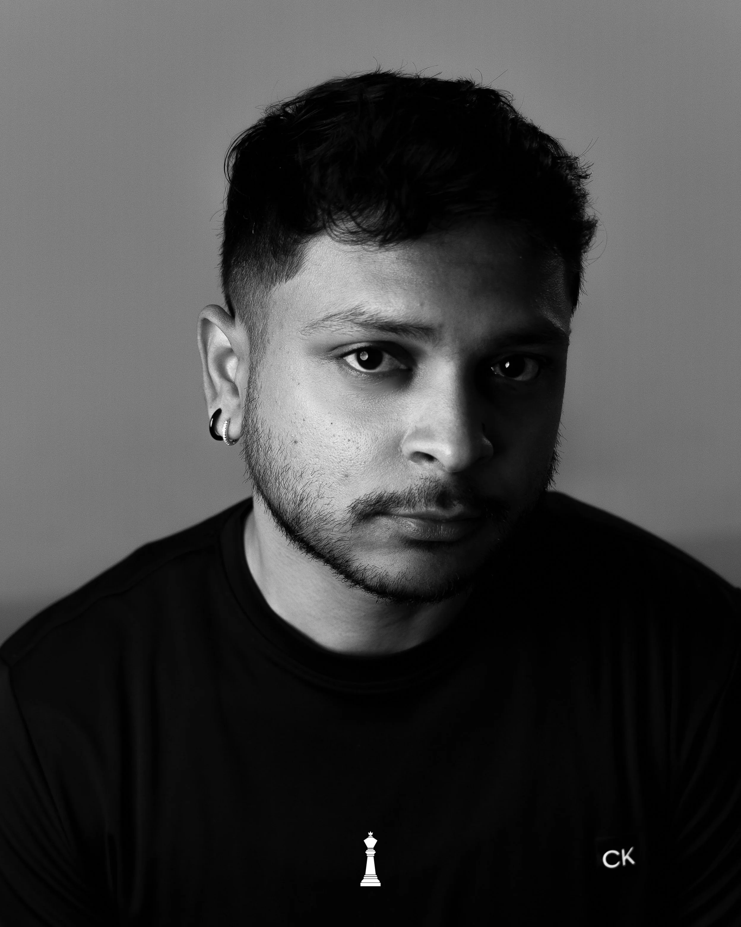 Black and white portrait of a young man with short dark hair, earrings, a textured beard, wearing a dark shirt with a small chess piece logo and the initials 'CK'.