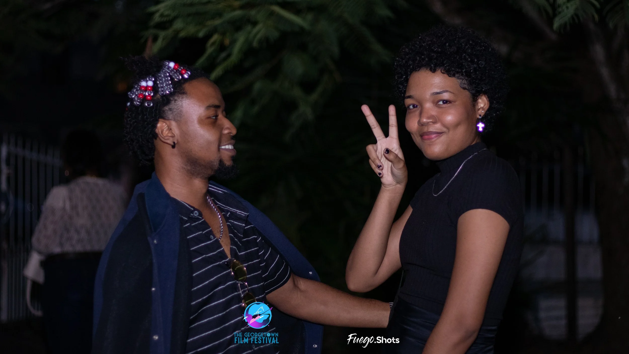Two young women with dark curly hair, one making a peace sign and both smiling, standing outdoors at night with dark green foliage in the background.