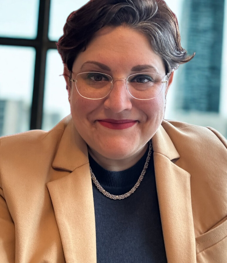A woman with short dark hair, glasses, and red lipstick smiling at the camera, wearing a beige blazer, black top, and silver necklace, indoors with large windows and a cityscape in the background.