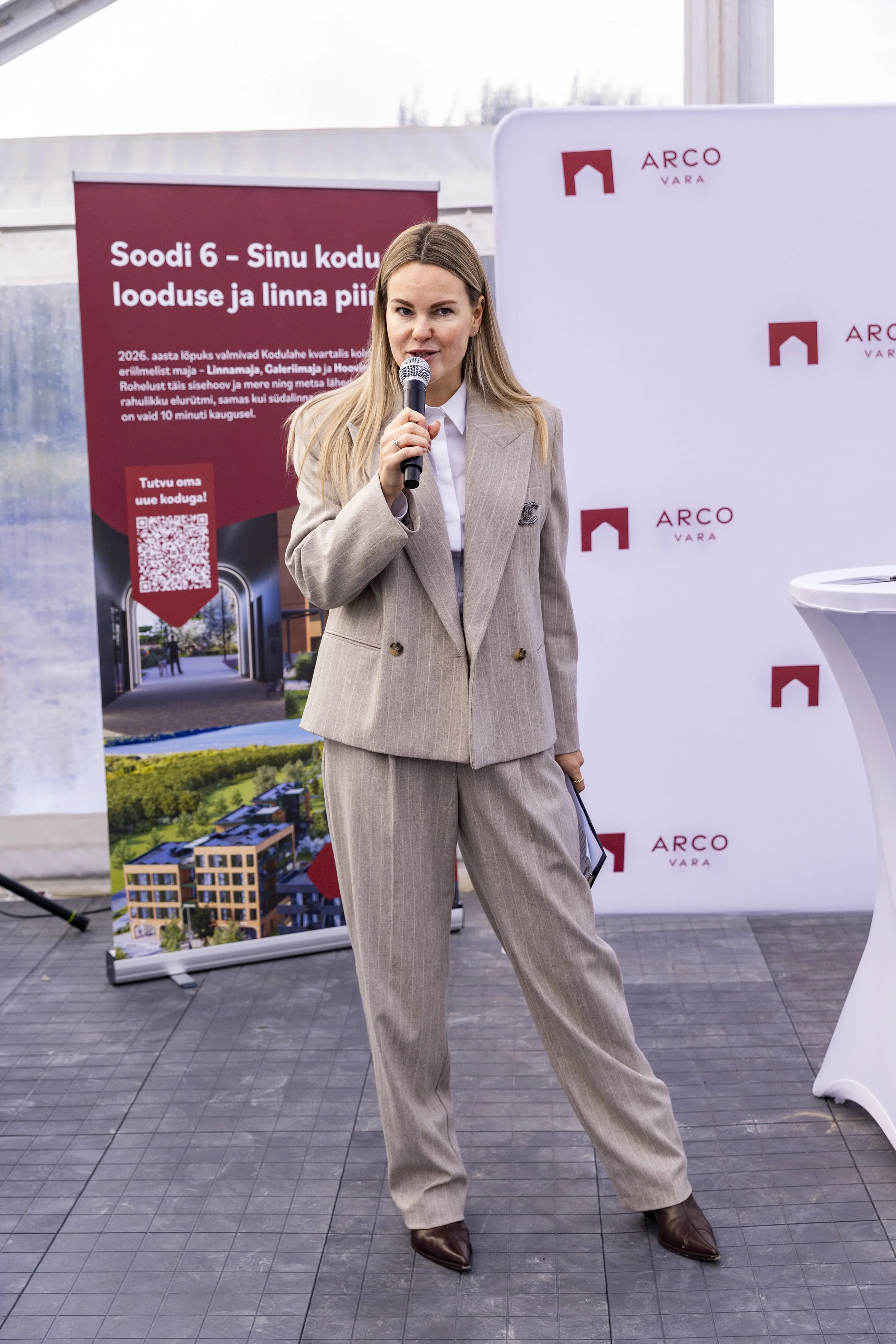 A woman in a tan striped suit holding a microphone at an outdoor event with banners that display the logo 'ARCO VARA' and a poster with construction information in Estonian.