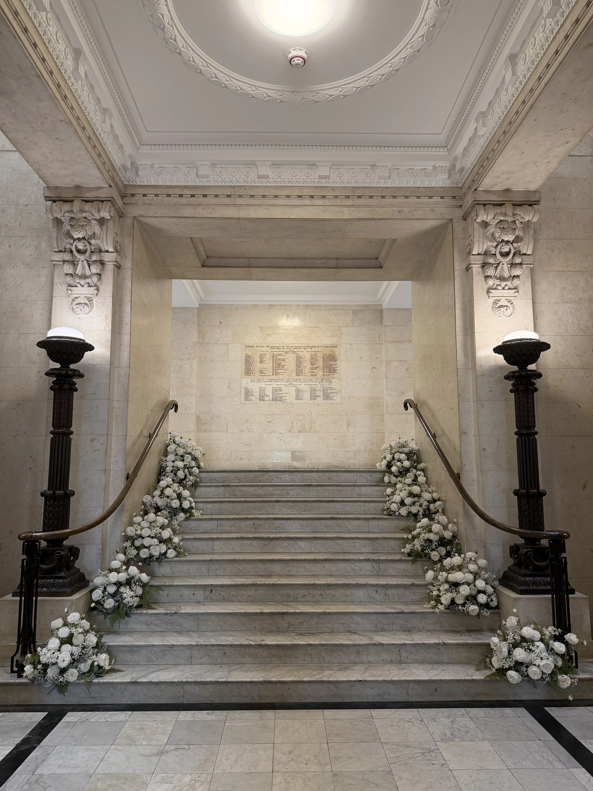 Marble staircase decorated with white floral arrangements on both sides, leading to an entrance in a grand building with ornate ceiling and wall details.