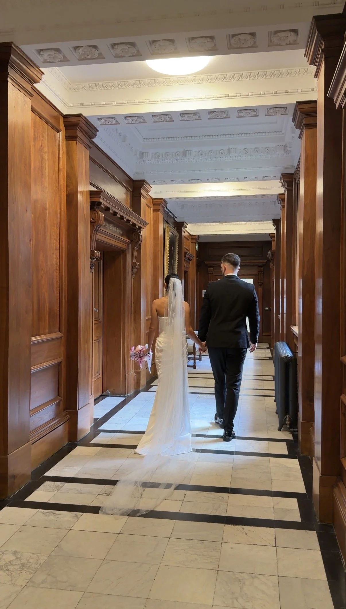 A bride and groom holding hands and walking down a hallway with wood-paneled walls, wearing wedding attire, the bride in a white gown with a veil holding a bouquet, and the groom in a dark suit.