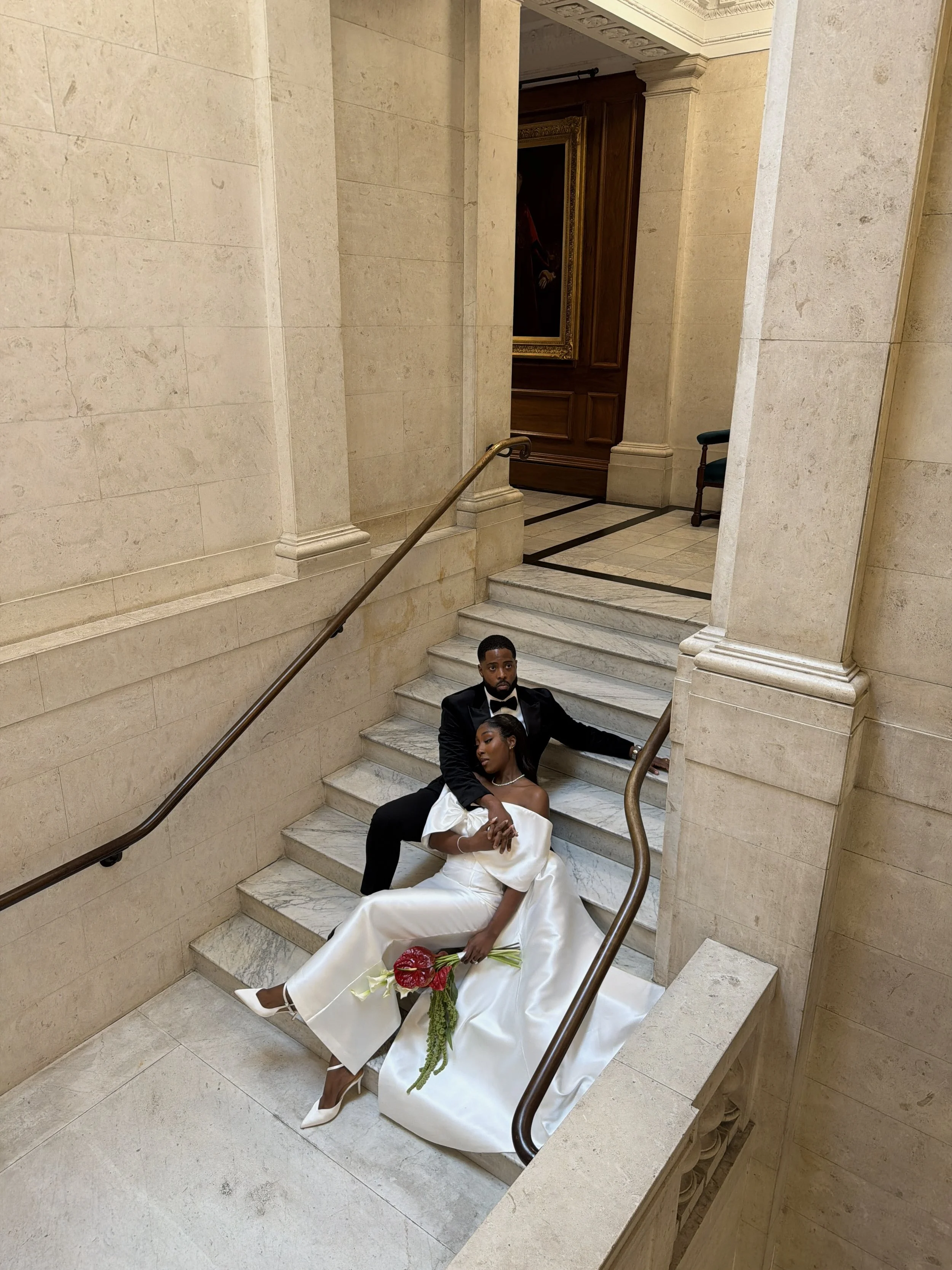A bride and groom sitting on a staircase in an elegant, historic building. The bride is wearing a white wedding dress and holding a bouquet of red flowers, while the groom is dressed in a black tuxedo with a bow tie.