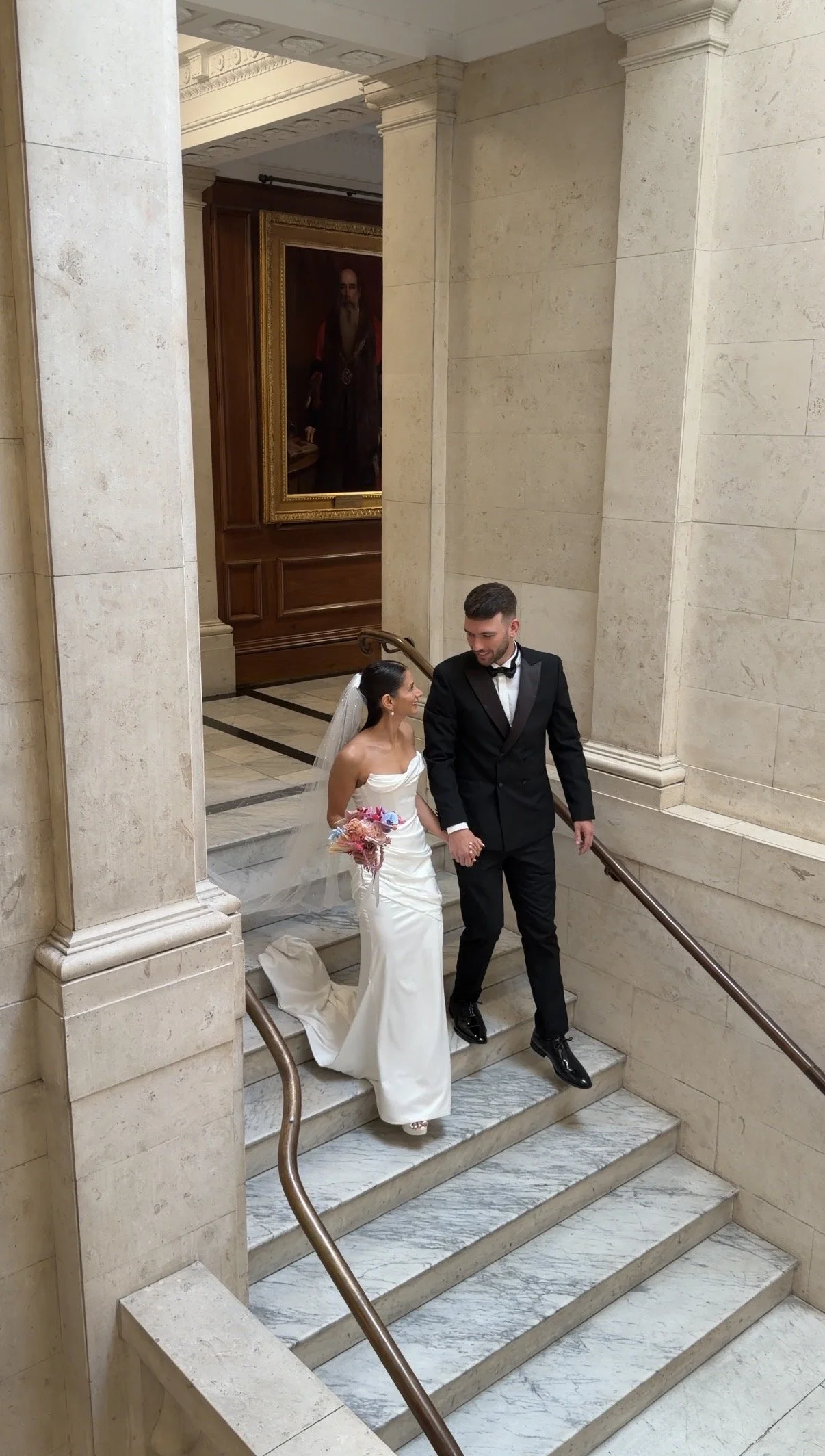 A bride and groom holding hands and descending marble stairs inside a grand building, with the bride in a white wedding dress holding a colorful bouquet, and the groom in a black tuxedo.