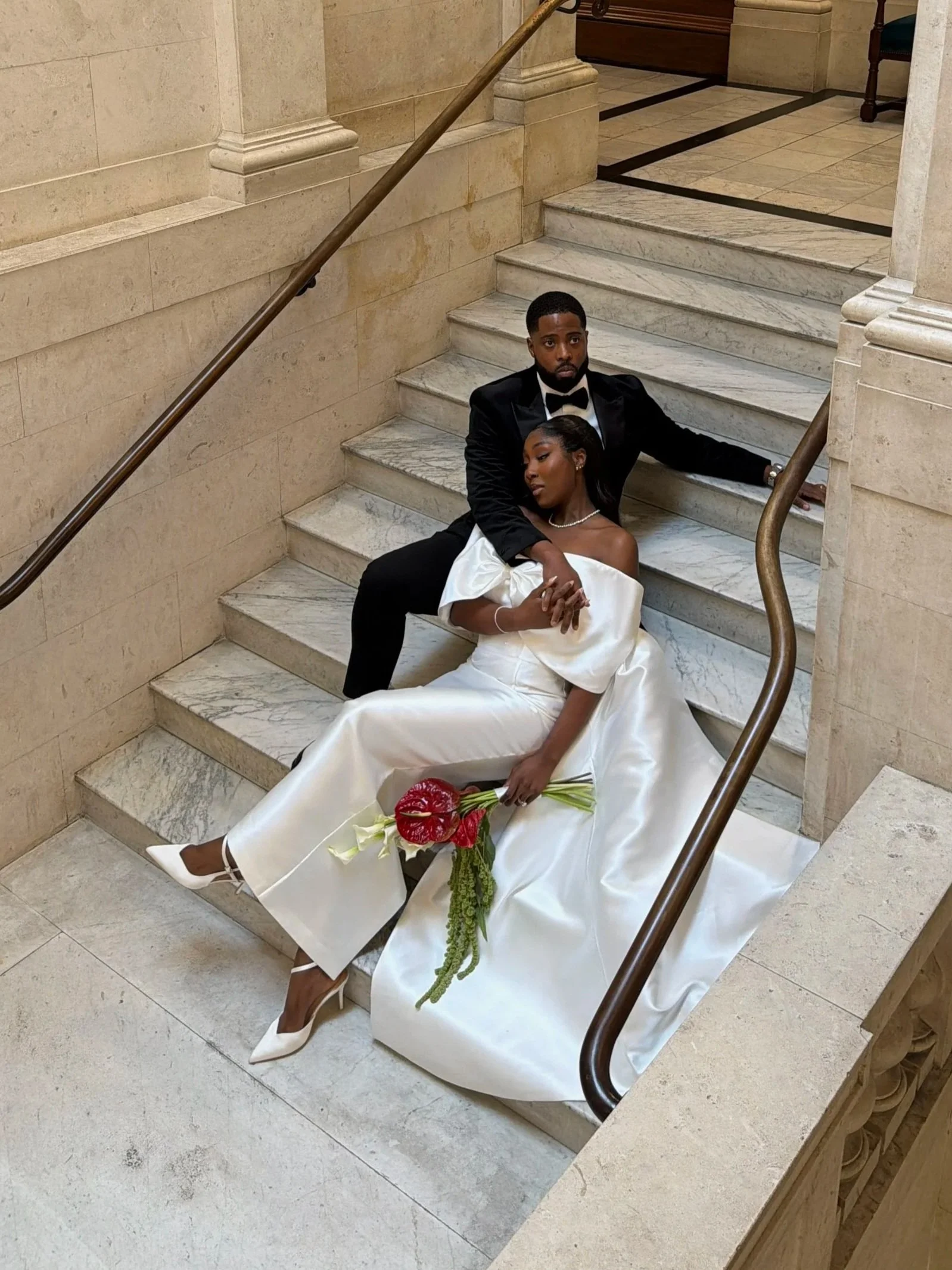 A man in a black tuxedo and a woman in a white wedding dress are sitting on marble stairs inside a building. The woman is holding a bouquet of red flowers and resting her head on the man's shoulder, while the man looks directly at the camera.