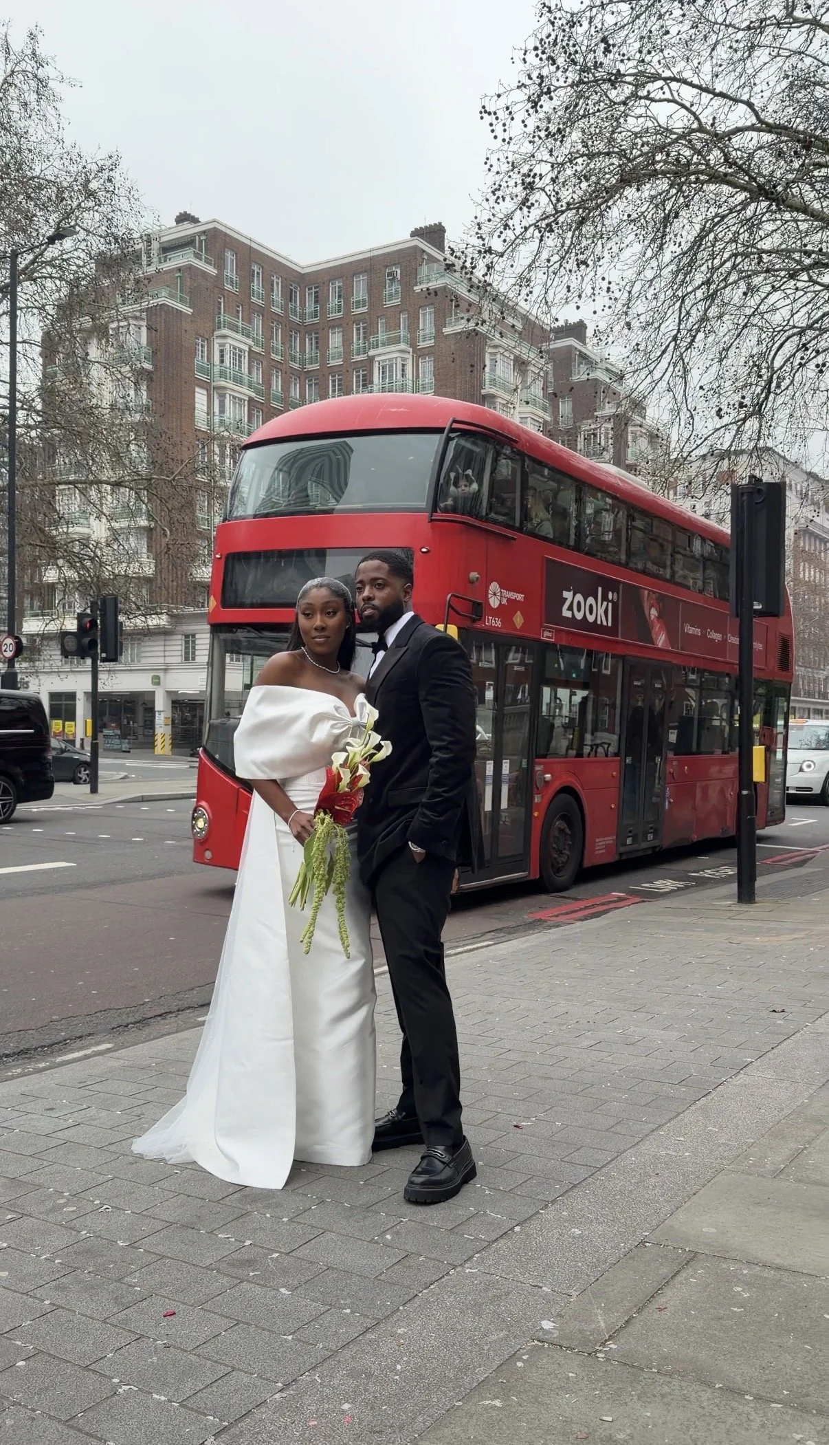 A bride and groom standing on a city sidewalk in front of a red double-decker bus, with buildings and trees in the background. The bride is wearing a white wedding dress and holding a bouquet, while the groom is dressed in a black suit and tie.