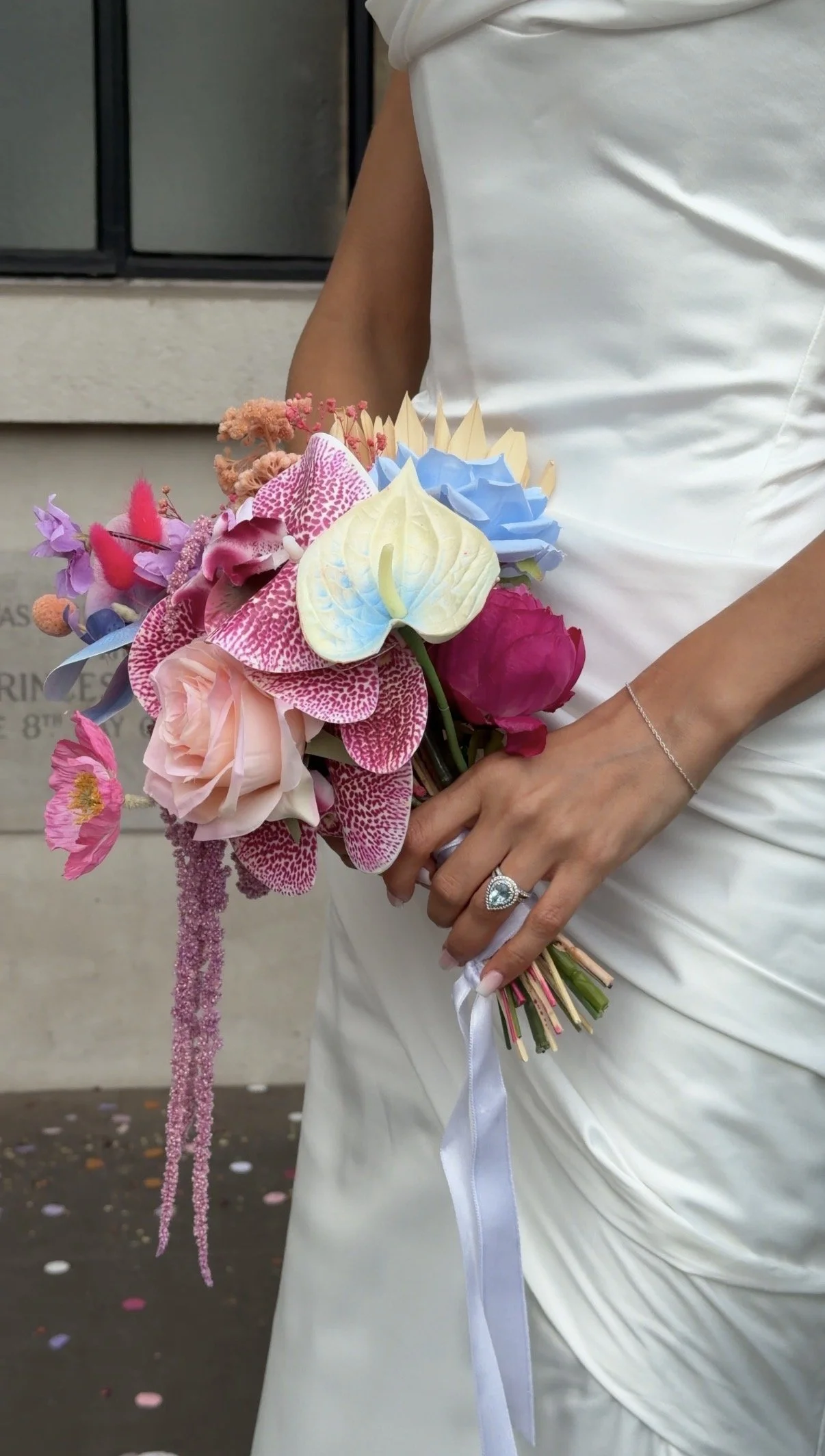 Person holding a colorful bouquet of flowers, including orchids, roses, and other blooms, while wearing a wedding dress and a ring.
