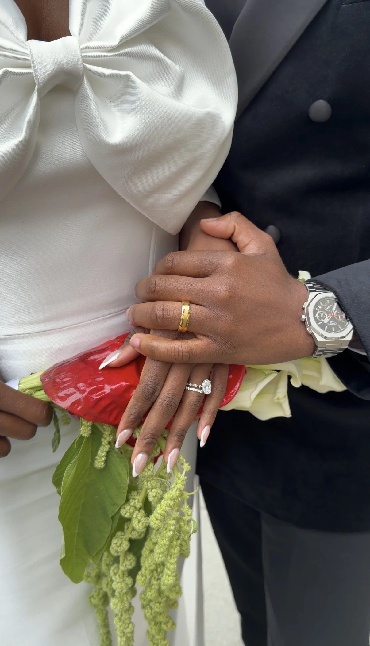 Close-up of a couple holding hands at a wedding, with the woman's hand displaying an engagement ring and a wedding band, and the man wearing a wristwatch. The bride's attire features white fabric with a bow, and both are holding a bouquet with green 