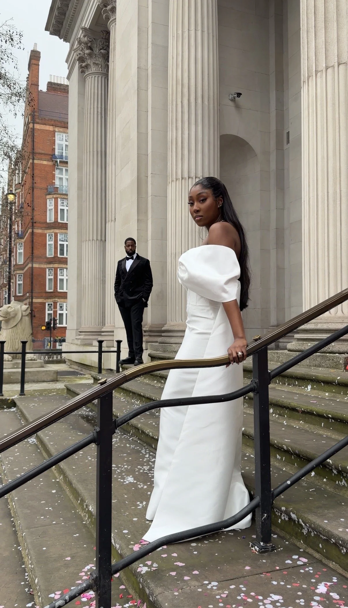 A woman in a white wedding dress with puffy sleeves standing on stairs outside a neoclassical building with tall columns, while a man in a black tuxedo stands in the background.