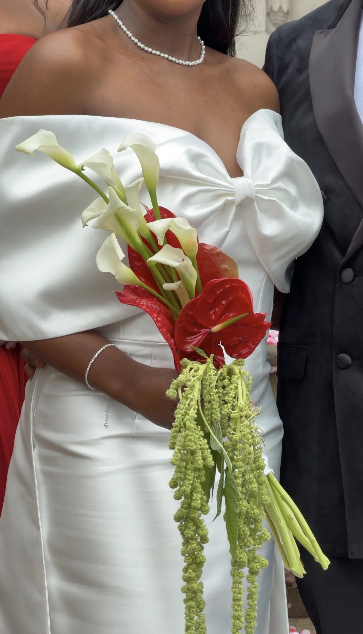 Close-up of a bride in a white dress holding a bouquet of white calla lilies, red anthuriums, and green hanging amaranthus, standing next to a groom in a dark suit at a wedding.