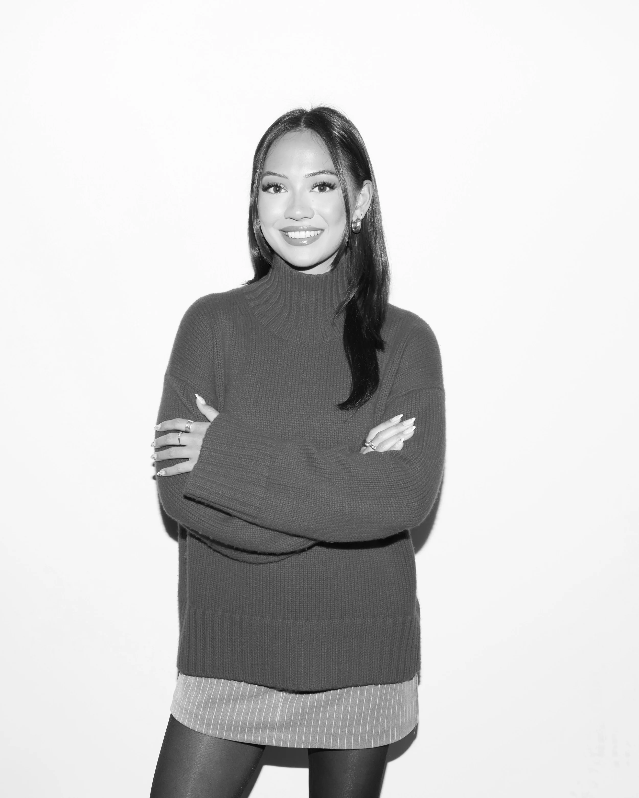 Black and white photo of a woman smiling with arms crossed, wearing a turtleneck sweater and a skirt.