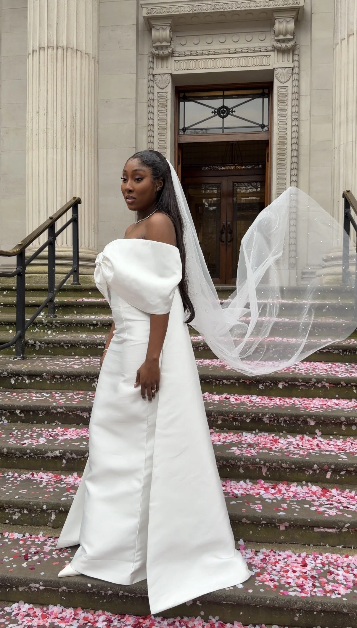 A bride in a white wedding gown with puffed sleeves stands on the steps of a historic building with columns, holding her long veil. Pink and white flower petals are scattered on the steps.