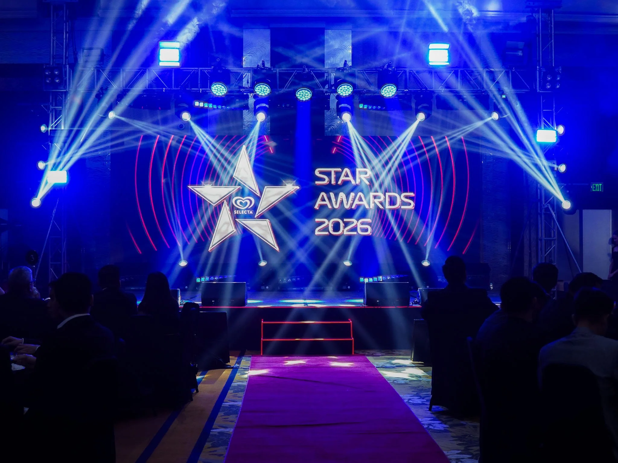 Stage with bright blue and white lighting, a large screen displaying a star logo and text 'Star Awards 2026,' and seated audience in a dark conference hall.