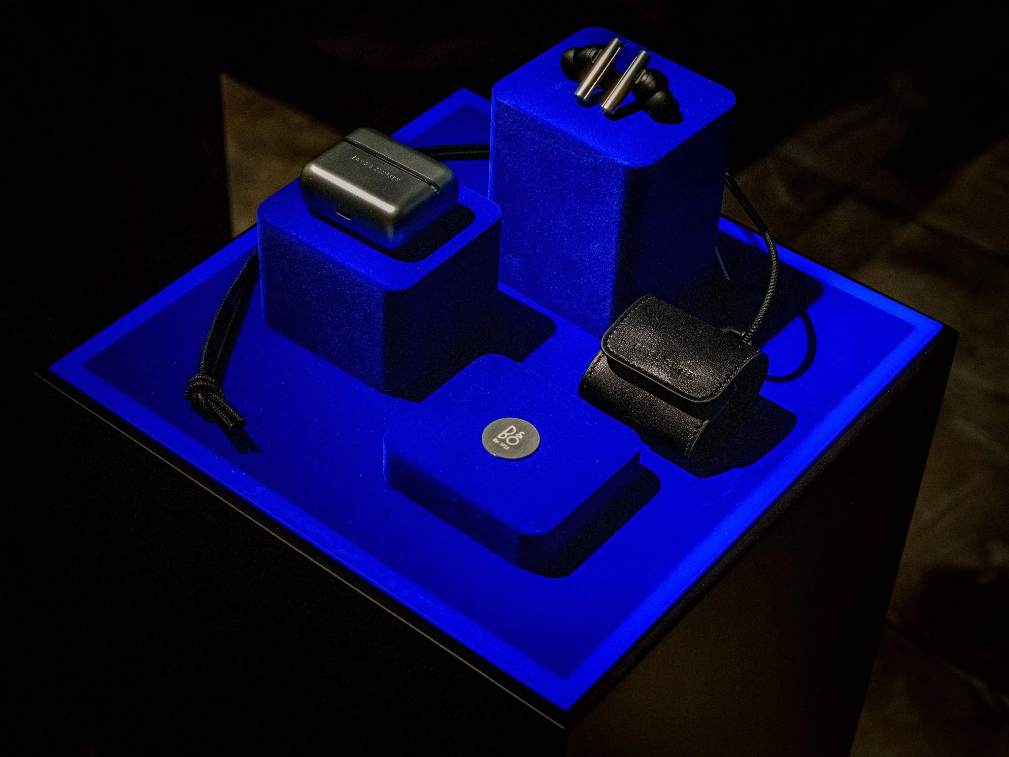 Display stand with black wireless earbuds, charger case, and leather case on blue-lit platform.