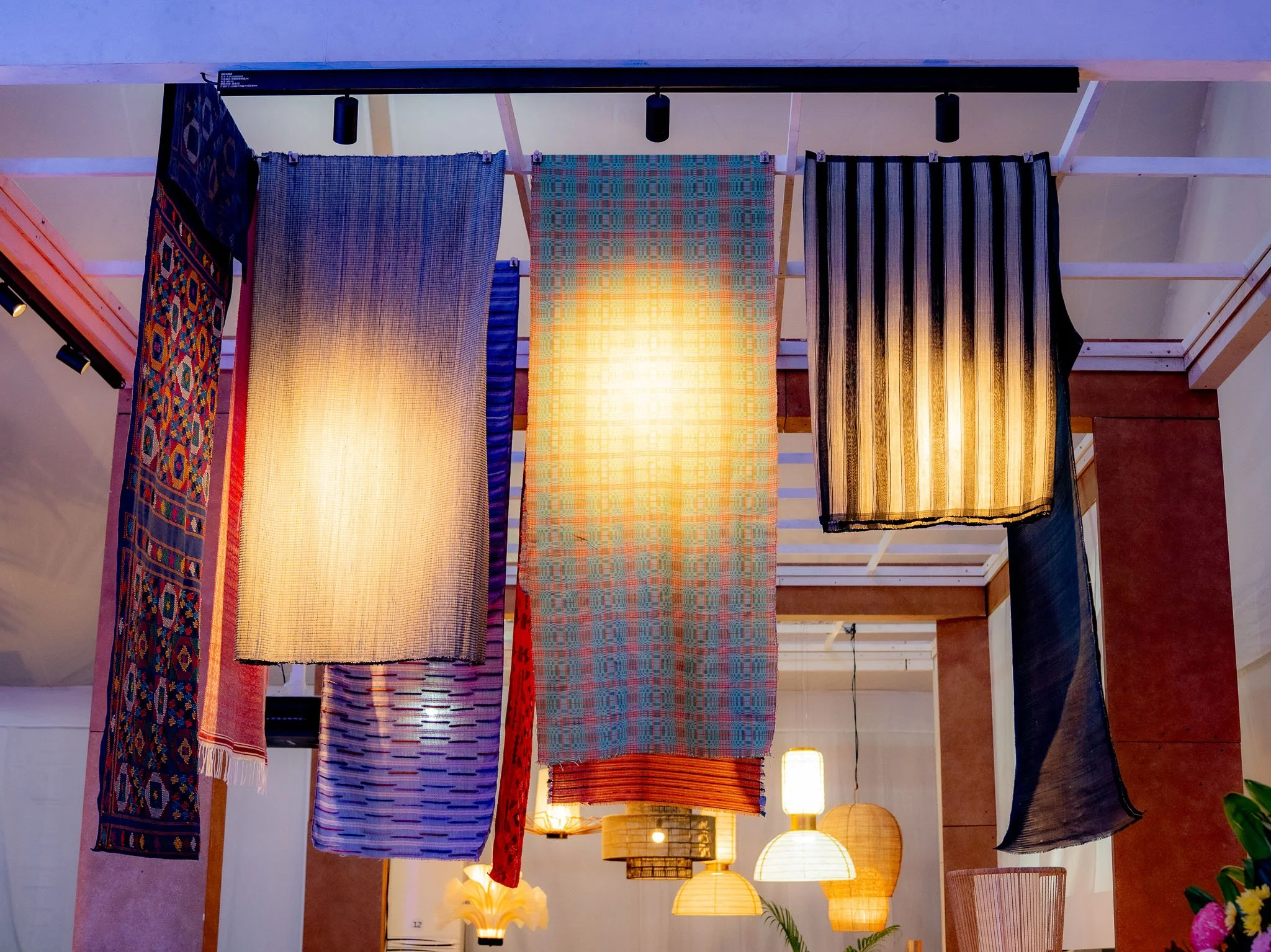 Display of various colorful textiles or scarves hanging from a ceiling rack in an indoor space with warm lighting.