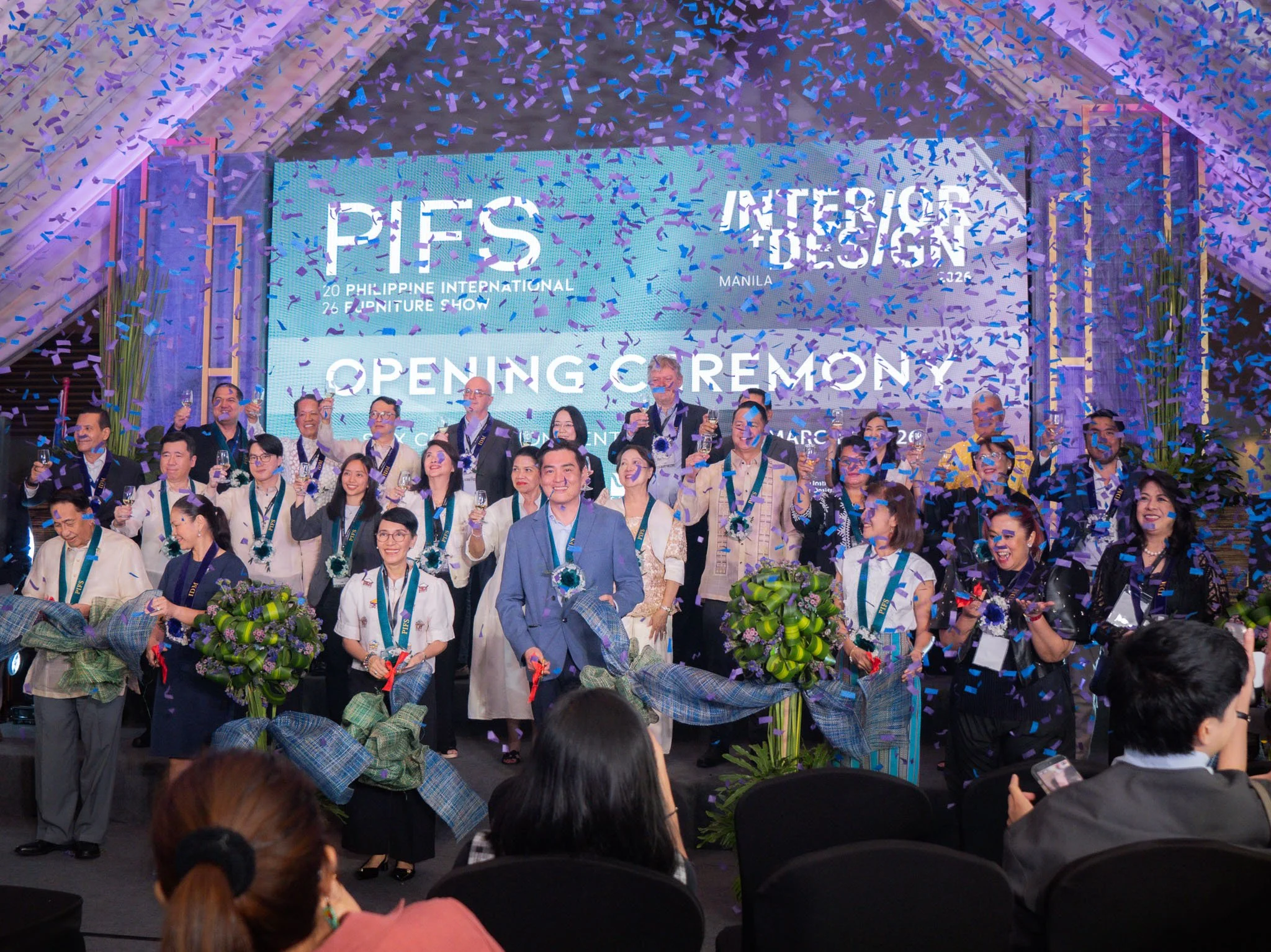 Group of people celebrating at an event with confetti, standing on stage with a large screen in the background displaying text about an interior design and furniture show, holding glasses in a toast.