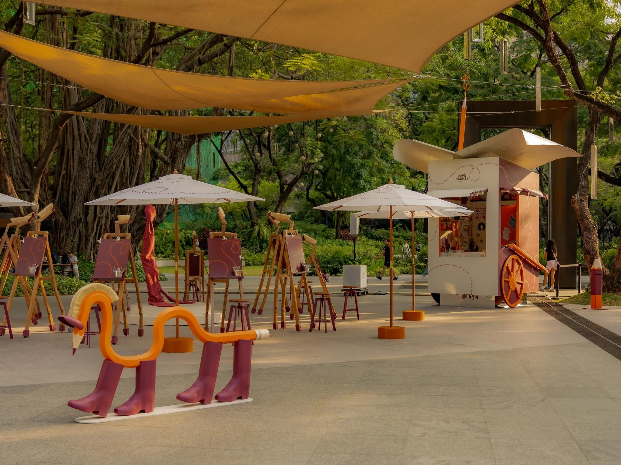 Outdoor art setup with easels, umbrellas, a whimsical shoe-shaped sculpture, and a white cart with a large paper top, surrounded by trees and shade fabric.