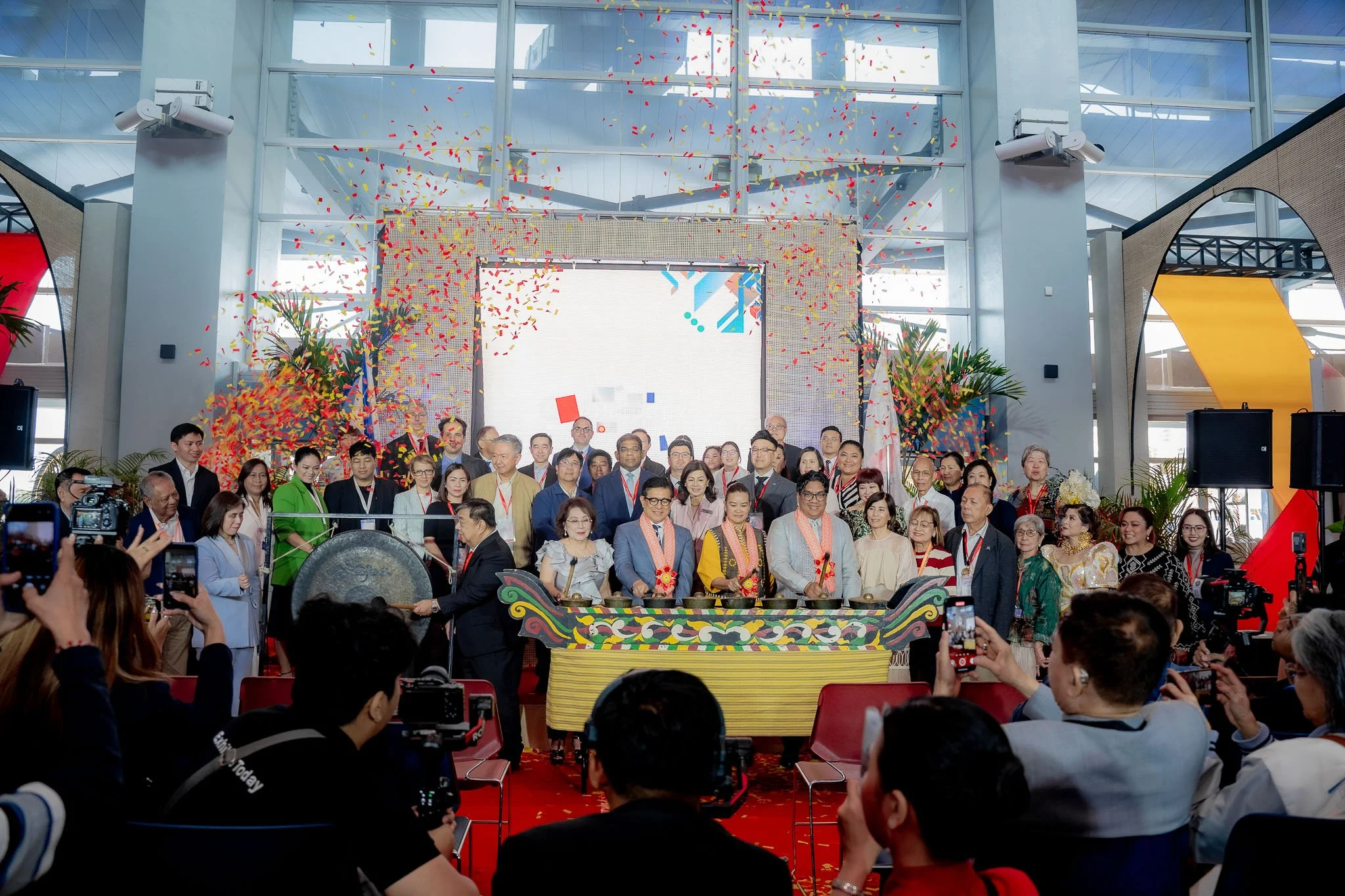 A large group of people gathered indoors for a celebration with confetti falling from above. They are standing behind a colorful traditional drum, and some individuals are taking pictures. The event is decorated with plants and a big digital screen in the background.